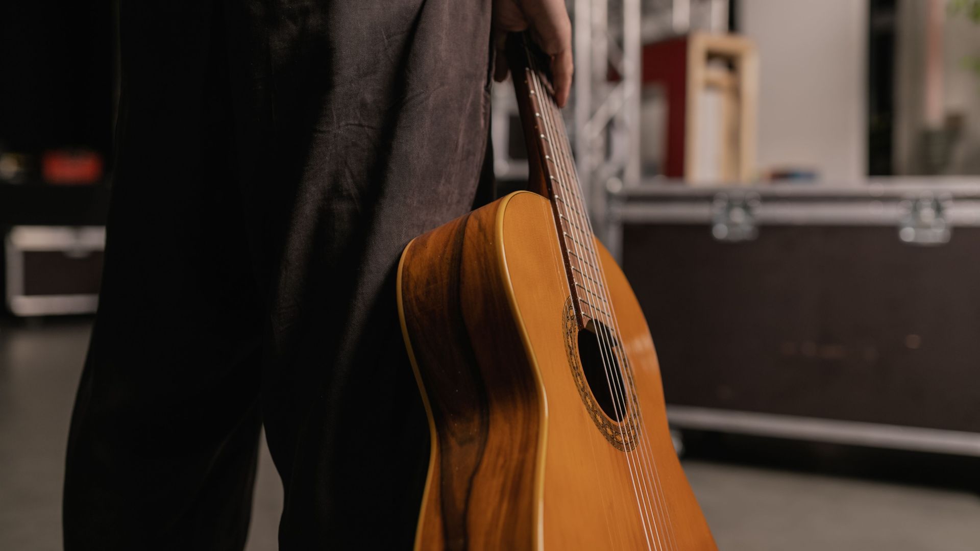 A person holding an acoustic guitar with a backstage setup in view, conveying a musical theme.