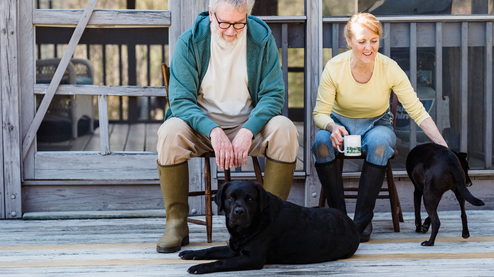 An elderly couple sits with their two dogs on a wooden porch, sharing a peaceful moment outdoors.