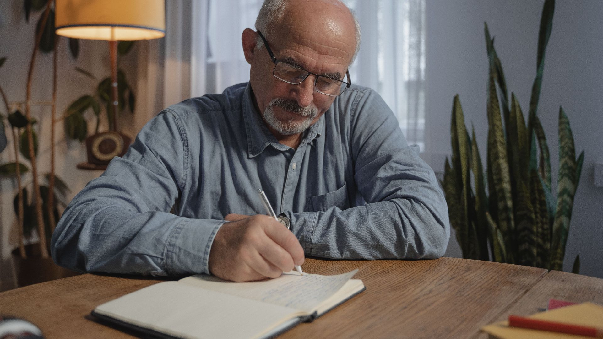 A senior adult man writing in a notebook at a desk in a cozy home office setting.