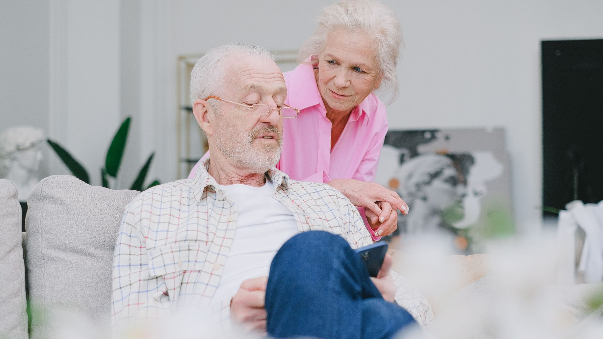 Elderly couple sitting indoors, engaged in a relaxed conversation while using a smartphone.