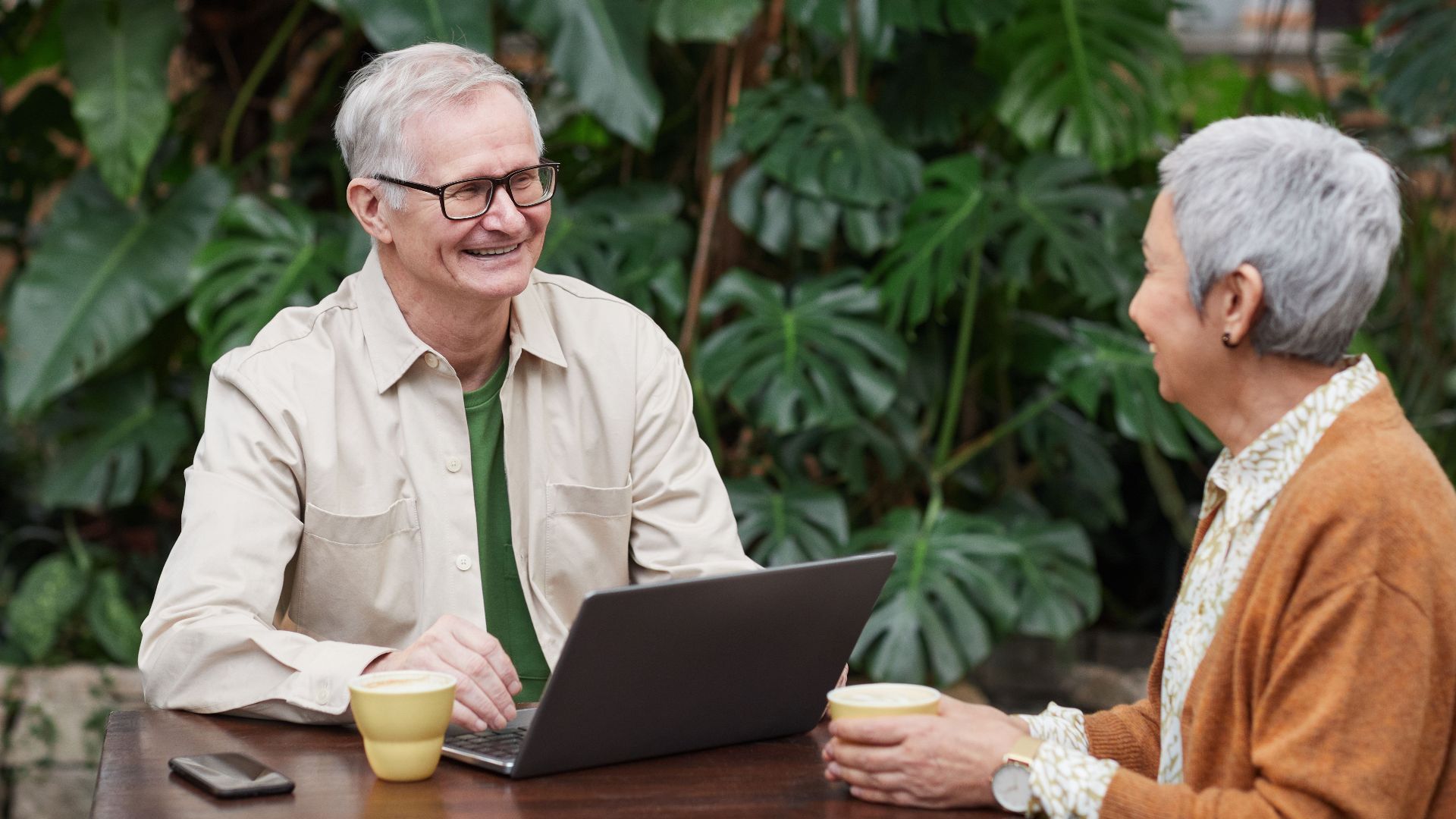 Senior couple smiling and enjoying coffee and conversation with laptop in a lush garden.