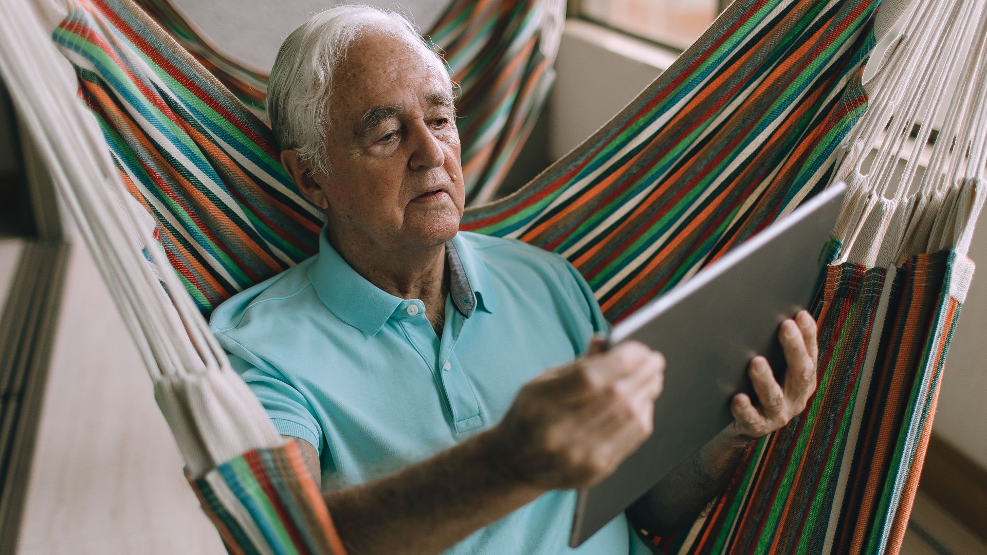 Elderly man using a tablet while relaxing on a hammock on a balcony.