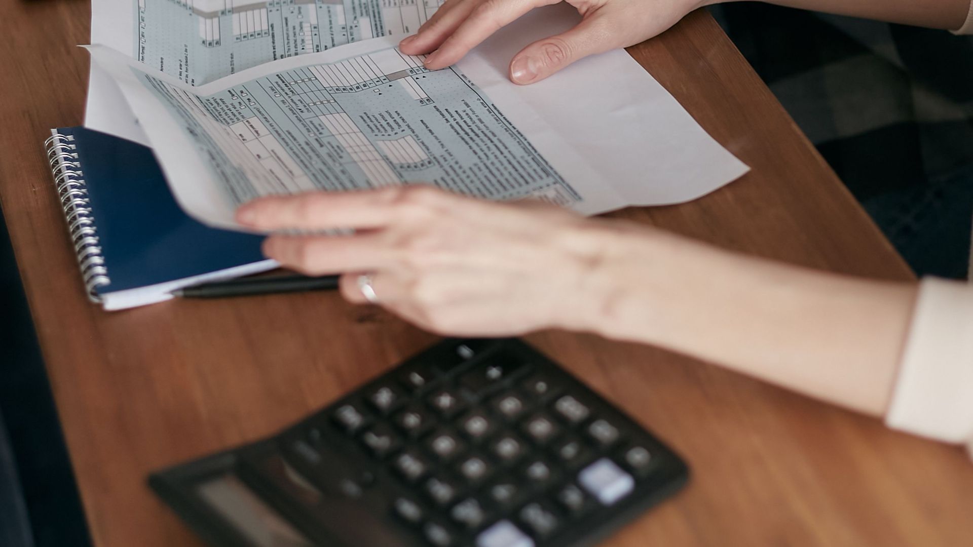 Close-up of hands organizing tax forms on a desk with a calculator, laptop, and notebook.