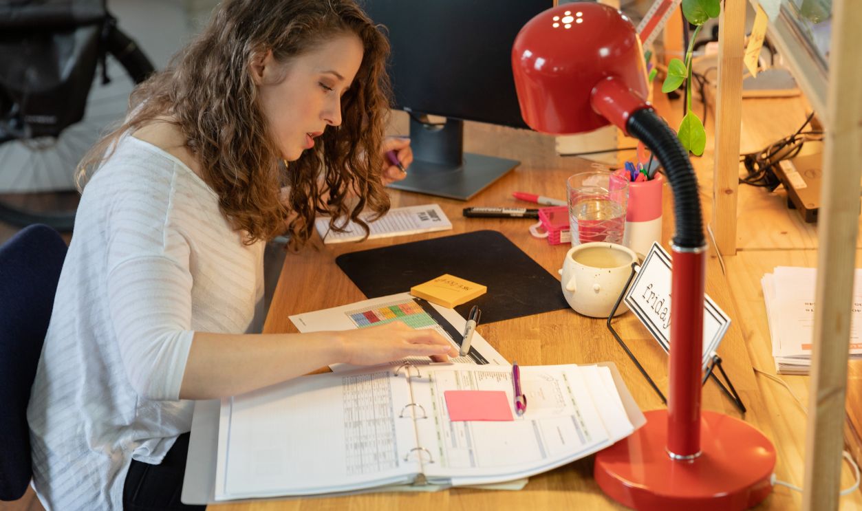 Woman working intently at her desk in a cozy office, surrounded by documents and a bright red lamp