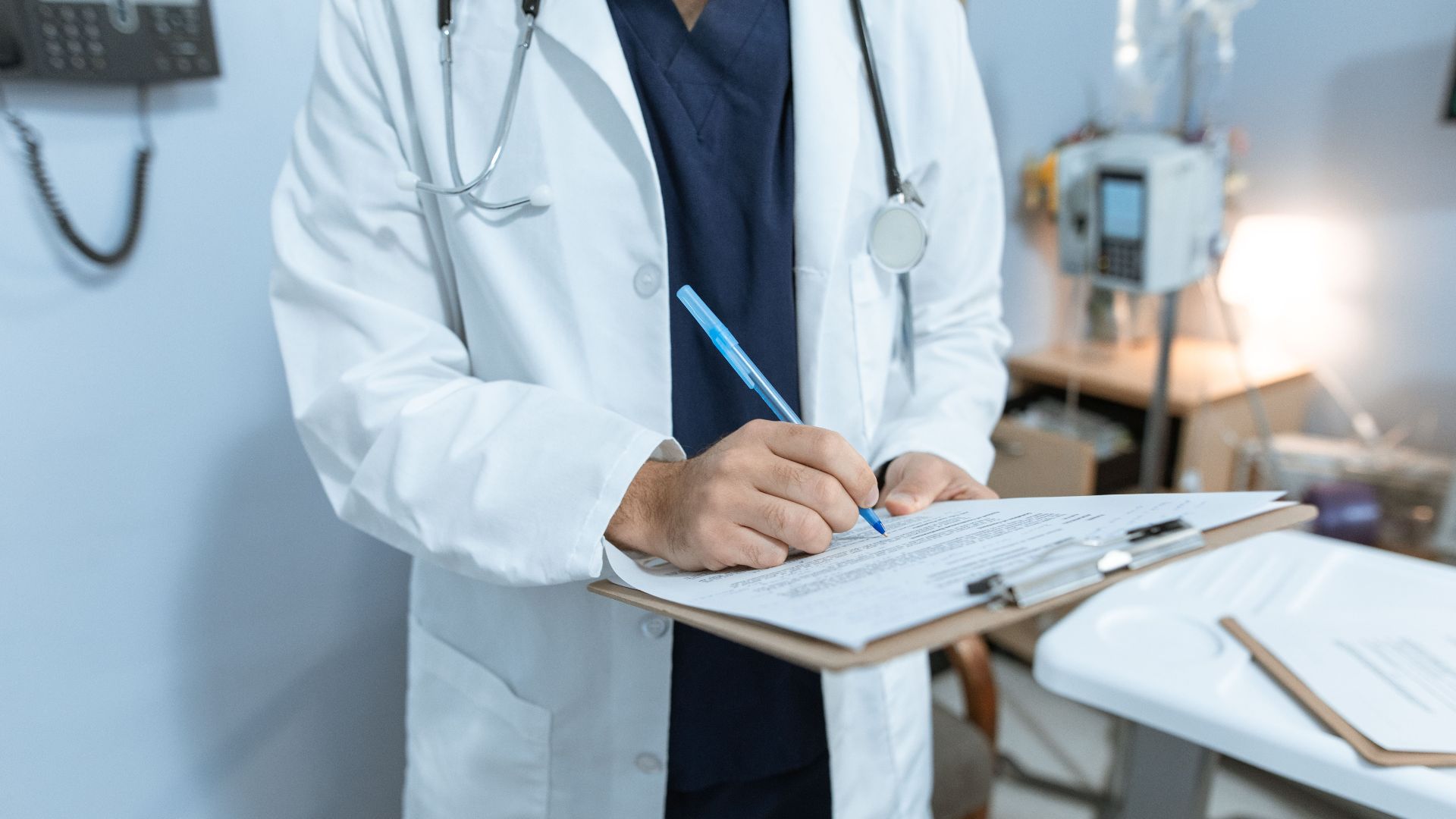 A doctor in a lab coat writing notes in a hospital setting, using a clipboard.