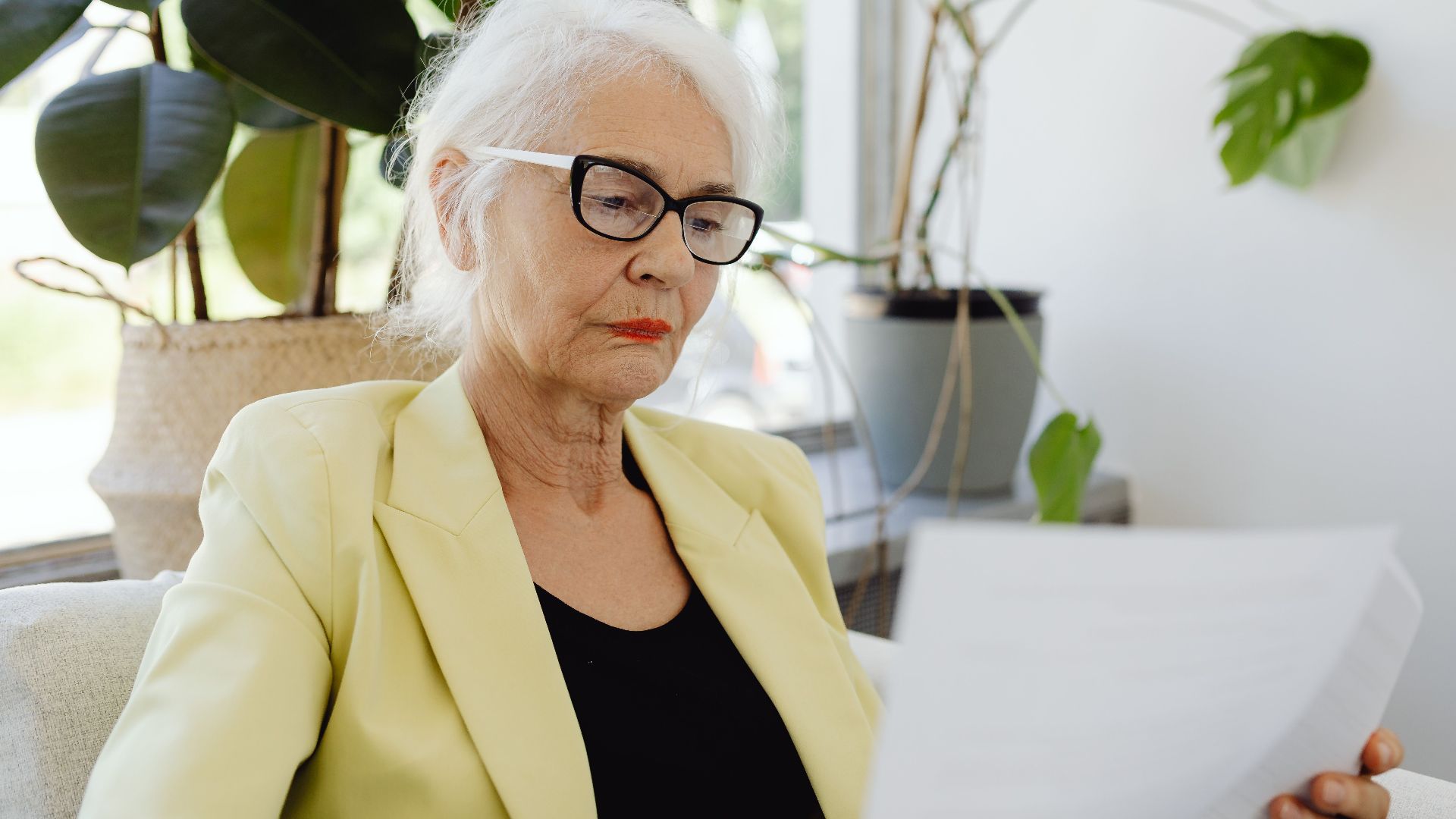 Confident senior woman in yellow jacket reading documents indoors.