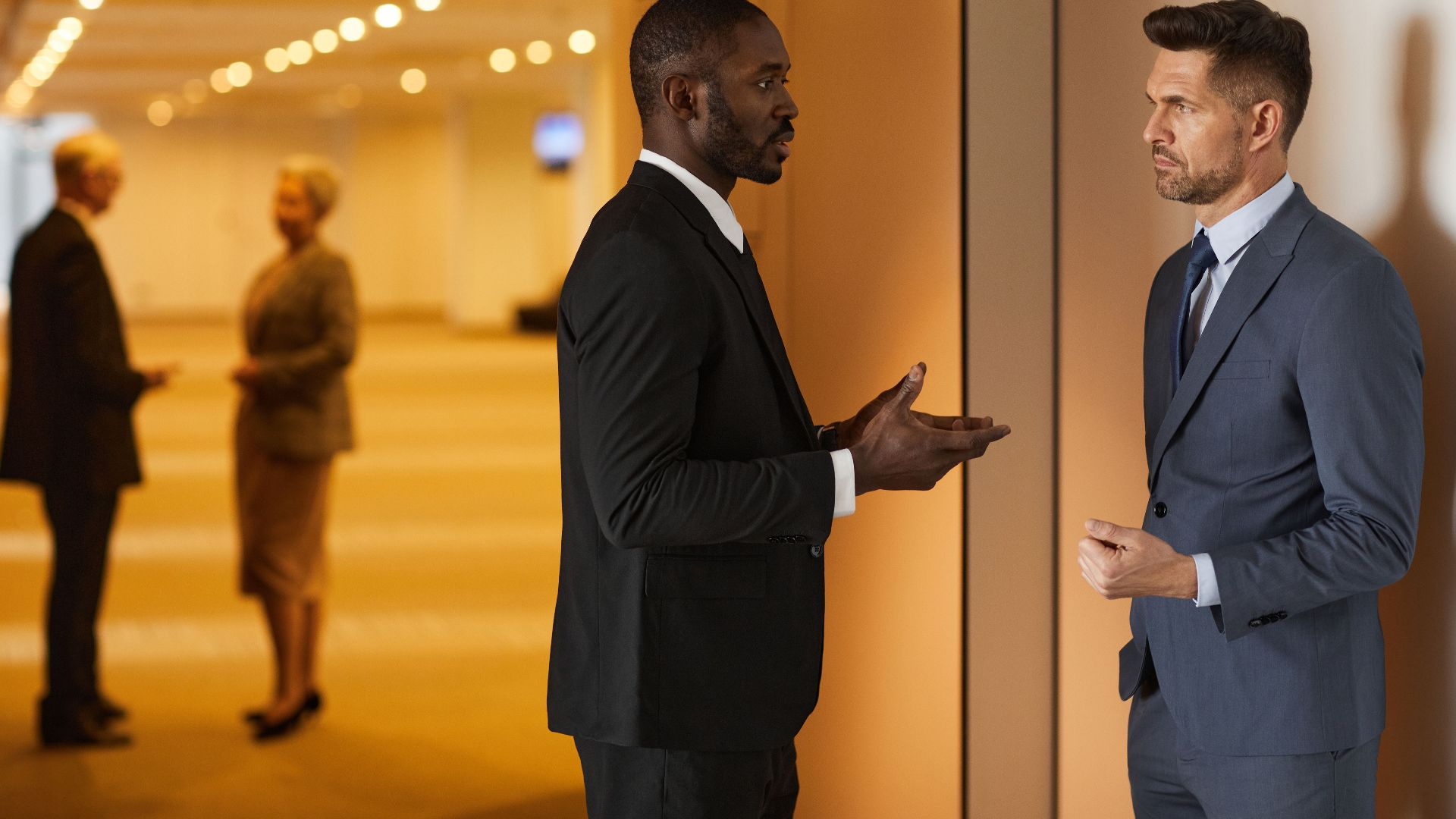 Two men in suits having a discussion in a modern hallway setting.