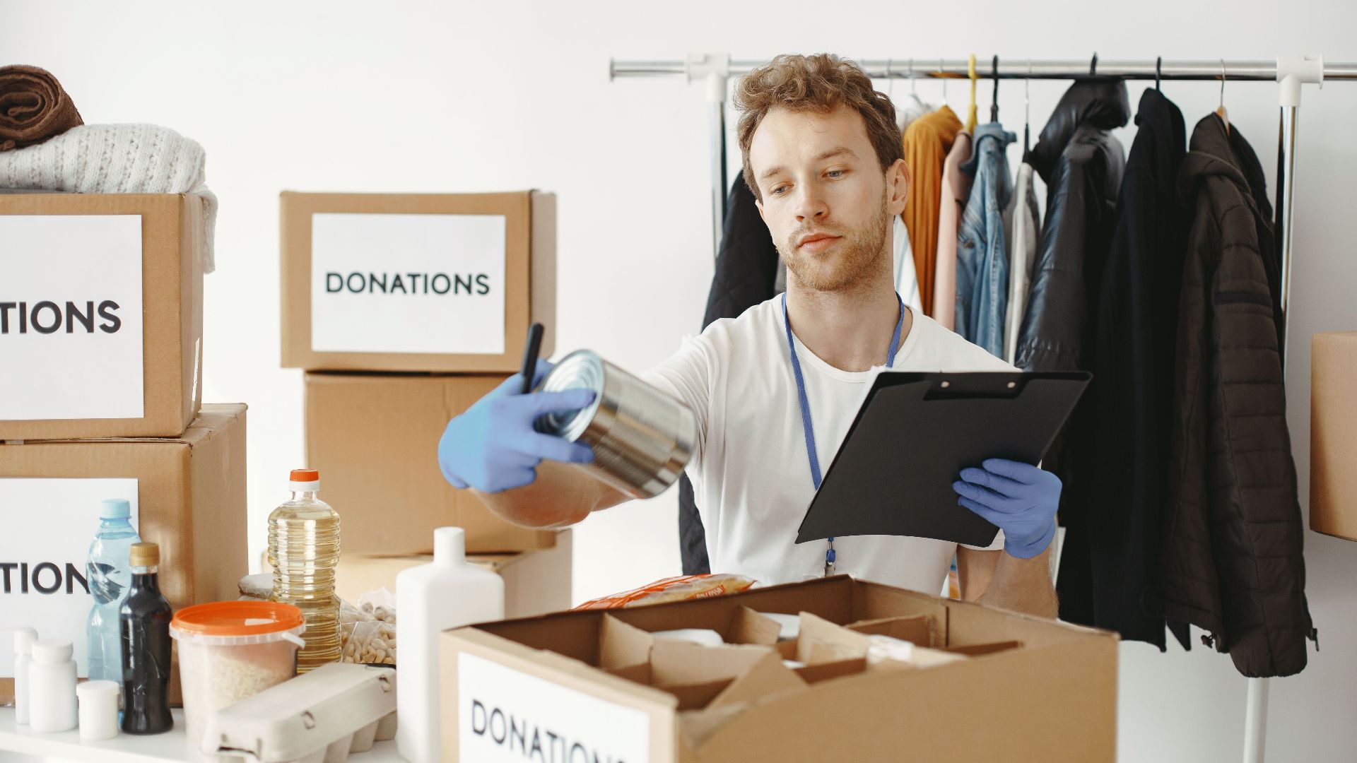 A volunteer organizes food and clothing donations in a community center, maintaining records on a clipboard.