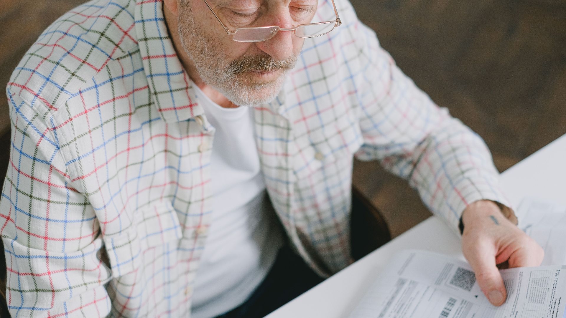 Elderly man in glasses carefully examines documents at home. Thoughtful and focused.