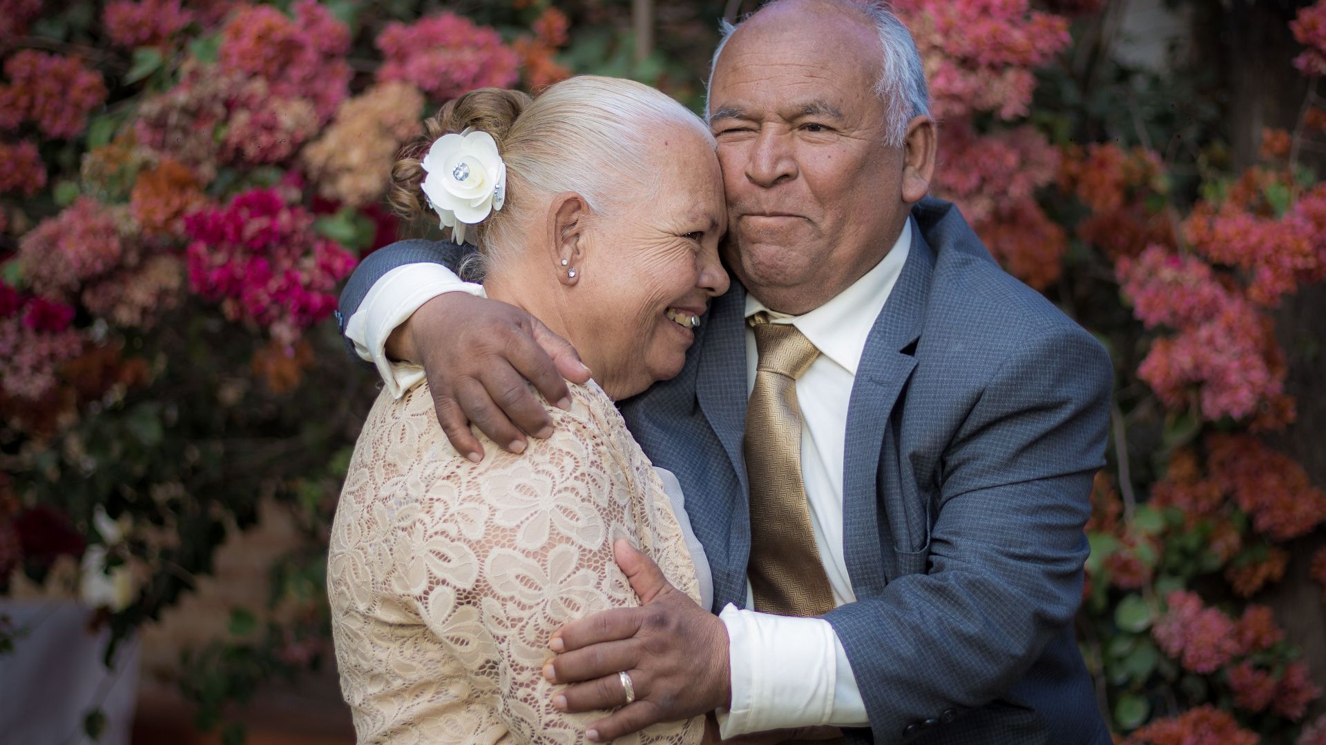 Smiling elderly couple in elegant attire embracing outdoors against a floral backdrop.