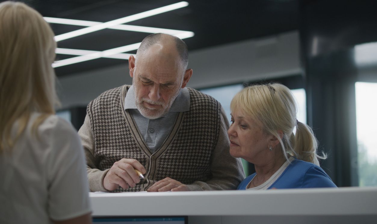 Elderly couple stand near reception desk in clinic lobby area, fill out papers. Female administrator talks to patients at information counter. Medical staff work in modern hospital or medical center.