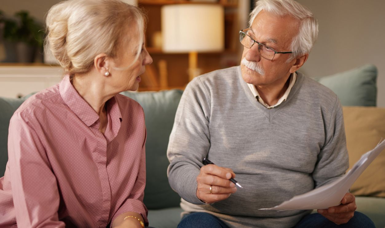 In the daytime, a senior couple sits on a sofa in their living room. The man reviews documents while the woman uses a calculator. They are seemingly discussing life insurance options.