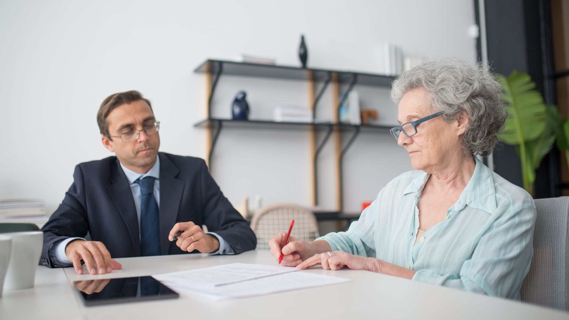 Elderly woman signing paperwork in modern office with consultant.