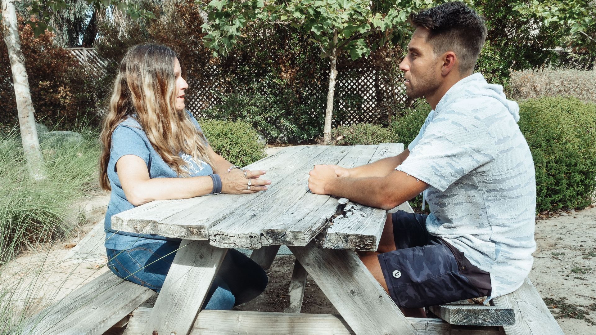 A mother and son sit talking at a rustic outdoor picnic table surrounded by greenery.