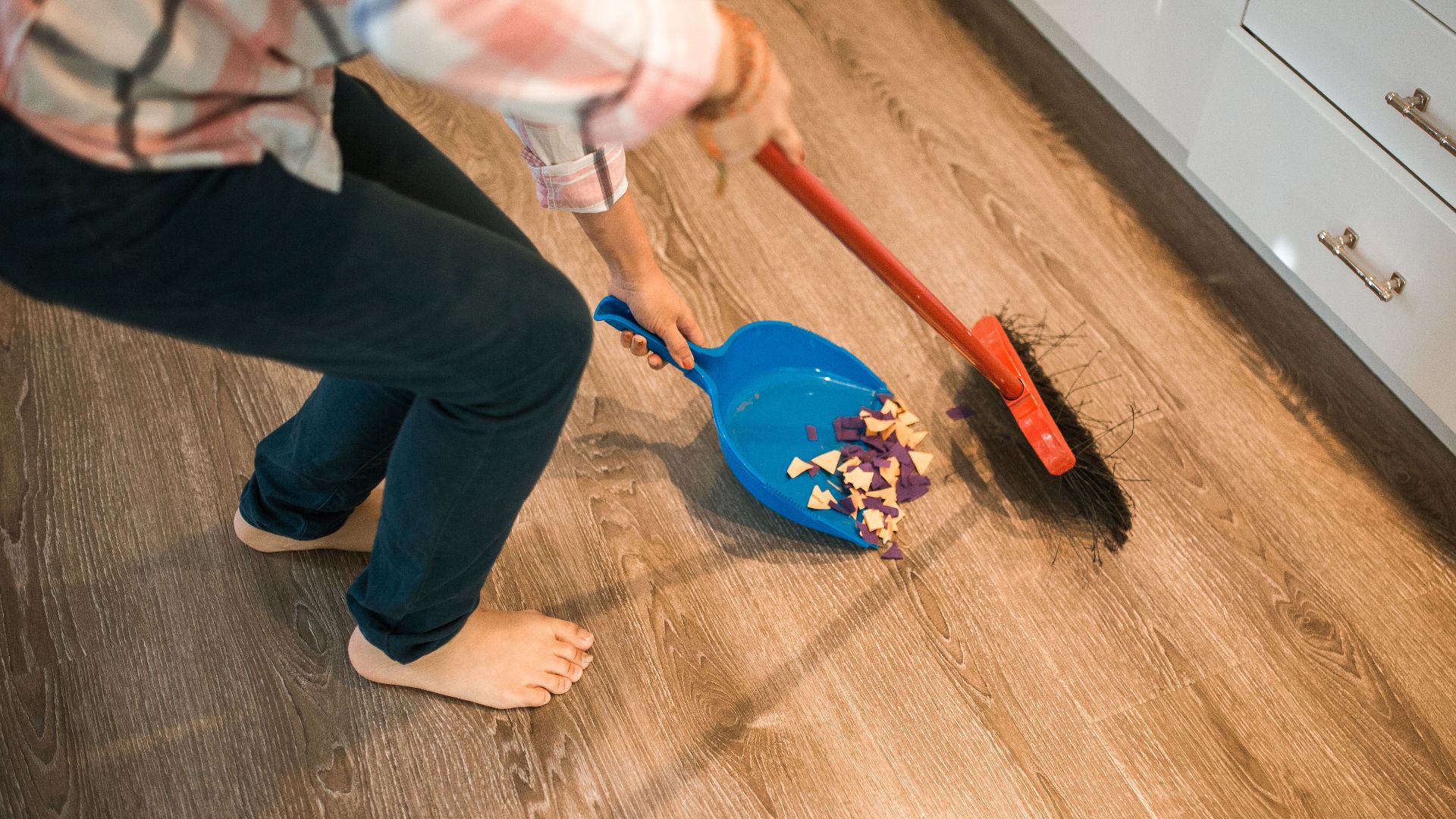 Close-up of a person sweeping debris into a blue dustpan on a wooden floor.