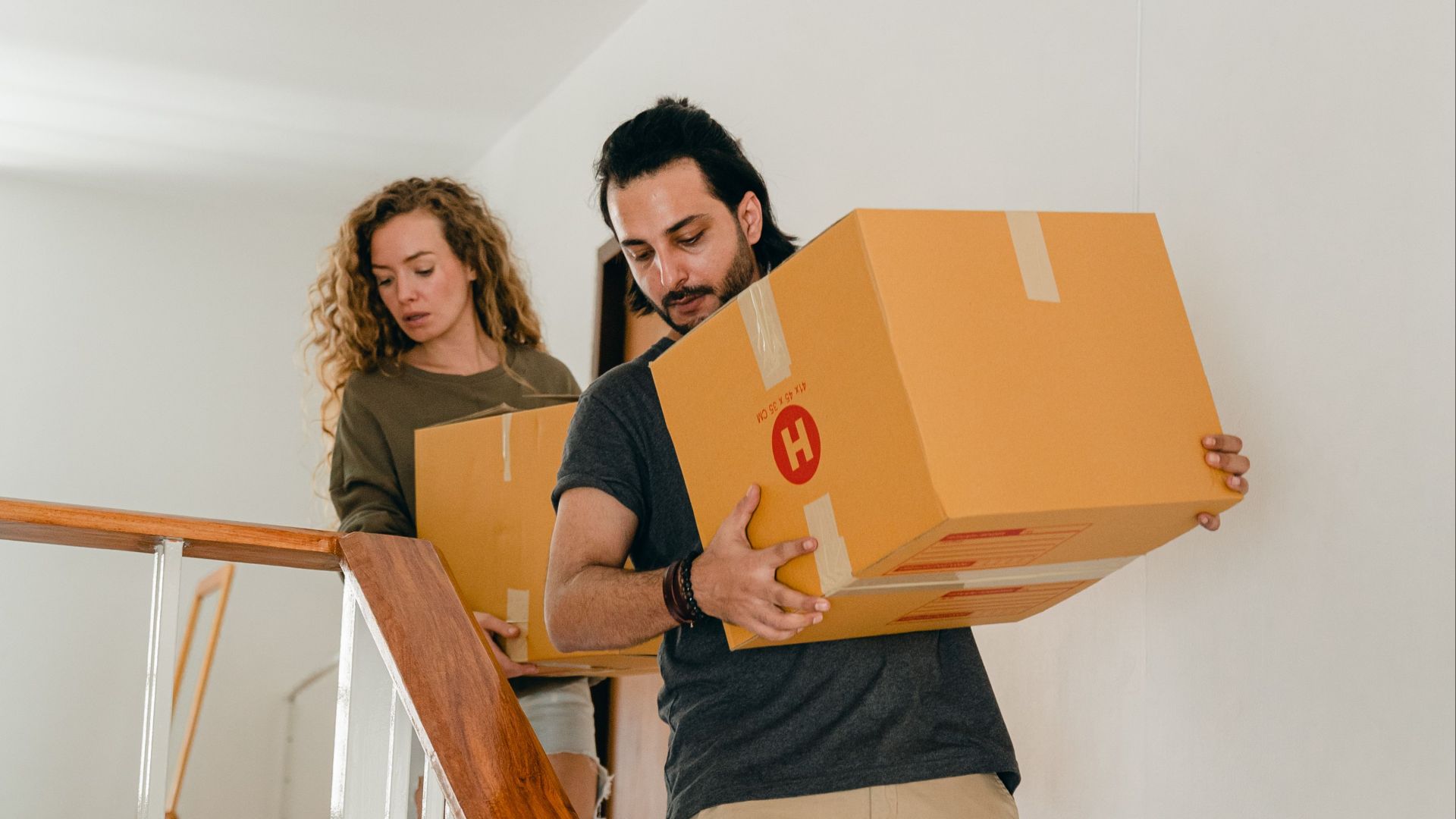 Full body of young woman and barefoot ethnic man in casual clothes carrying boxes on wooden stairs while moving into new home