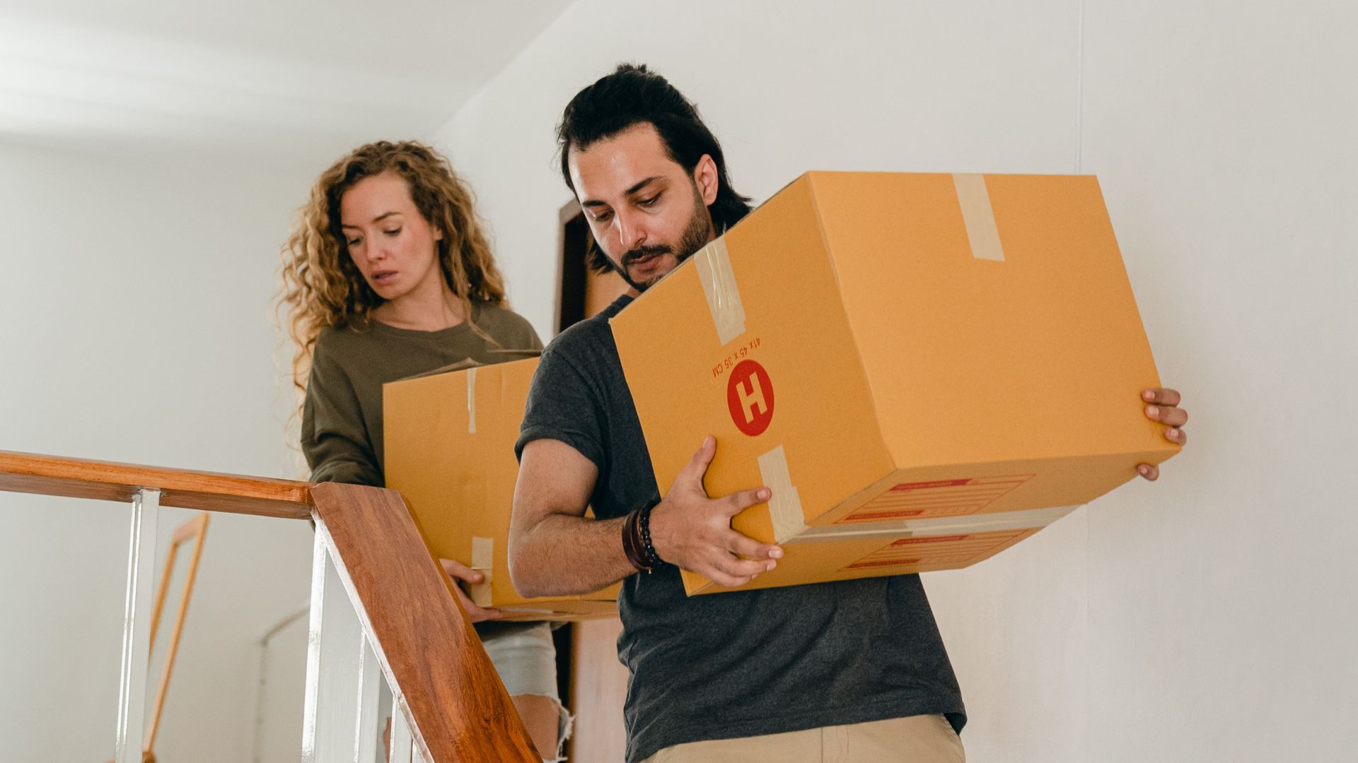 Full body of young woman and barefoot ethnic man in casual clothes carrying boxes on wooden stairs while moving into new home