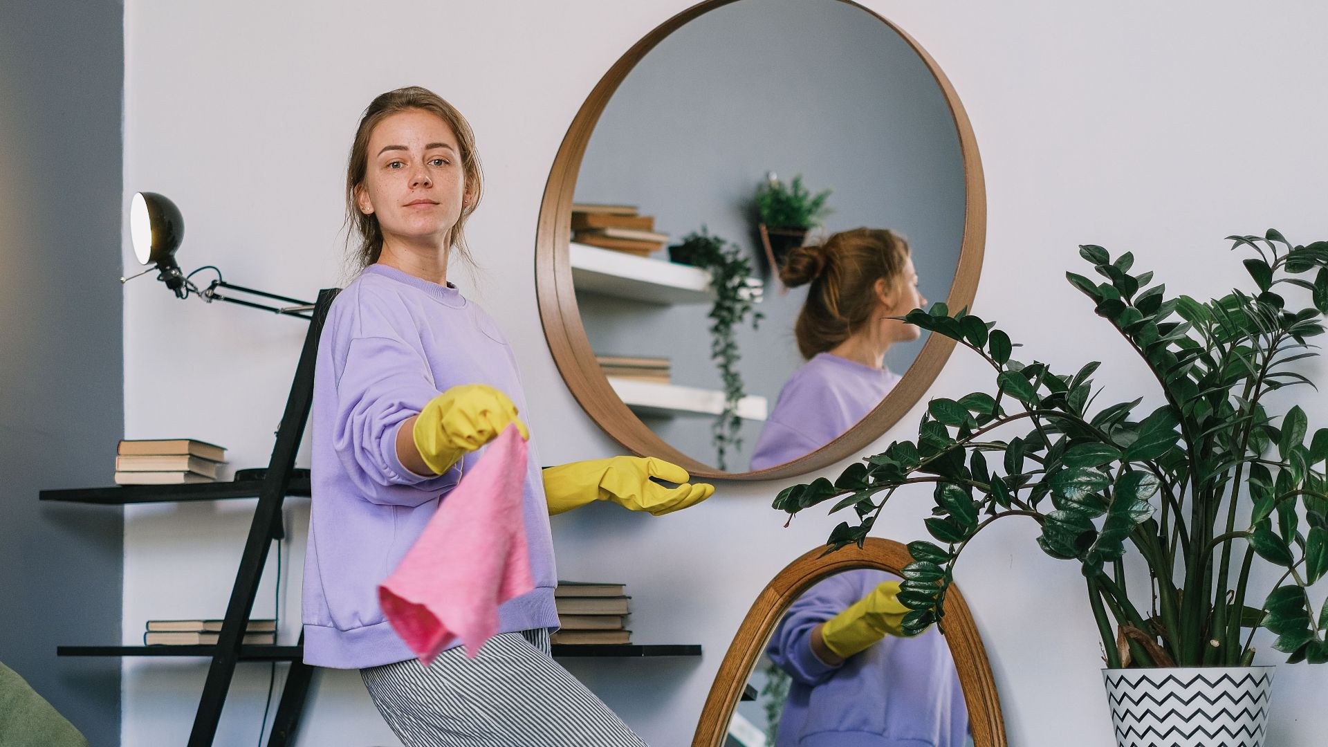 Side view of cheerful woman in casual clothes and latex gloves standing near mirrors while cleaning apartment in daytime