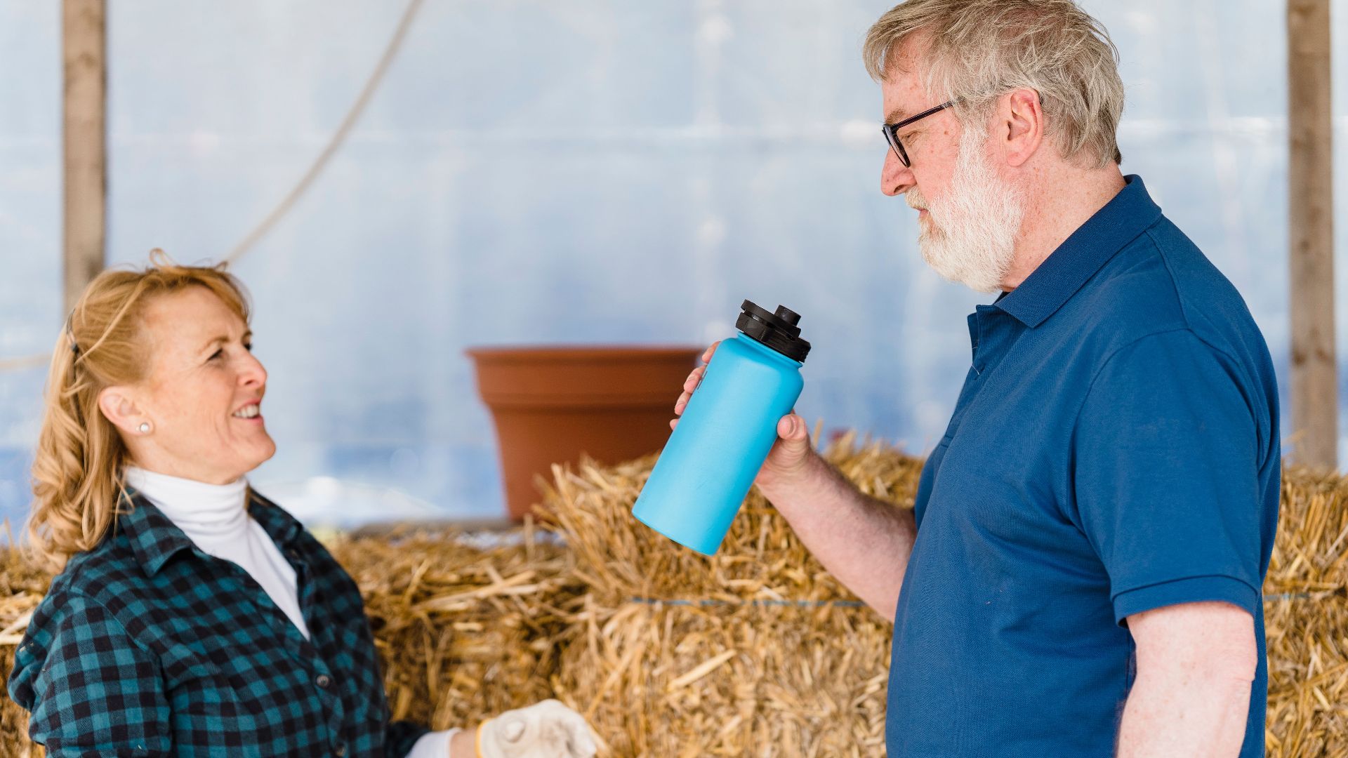 Elderly man and woman in a casual conversation indoors with a blue tumbler. Haystack in the background.