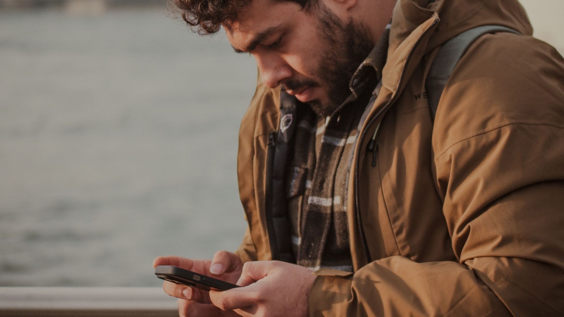 Man wearing a jacket, checking his smartphone by a waterfront, cityscape in the background.