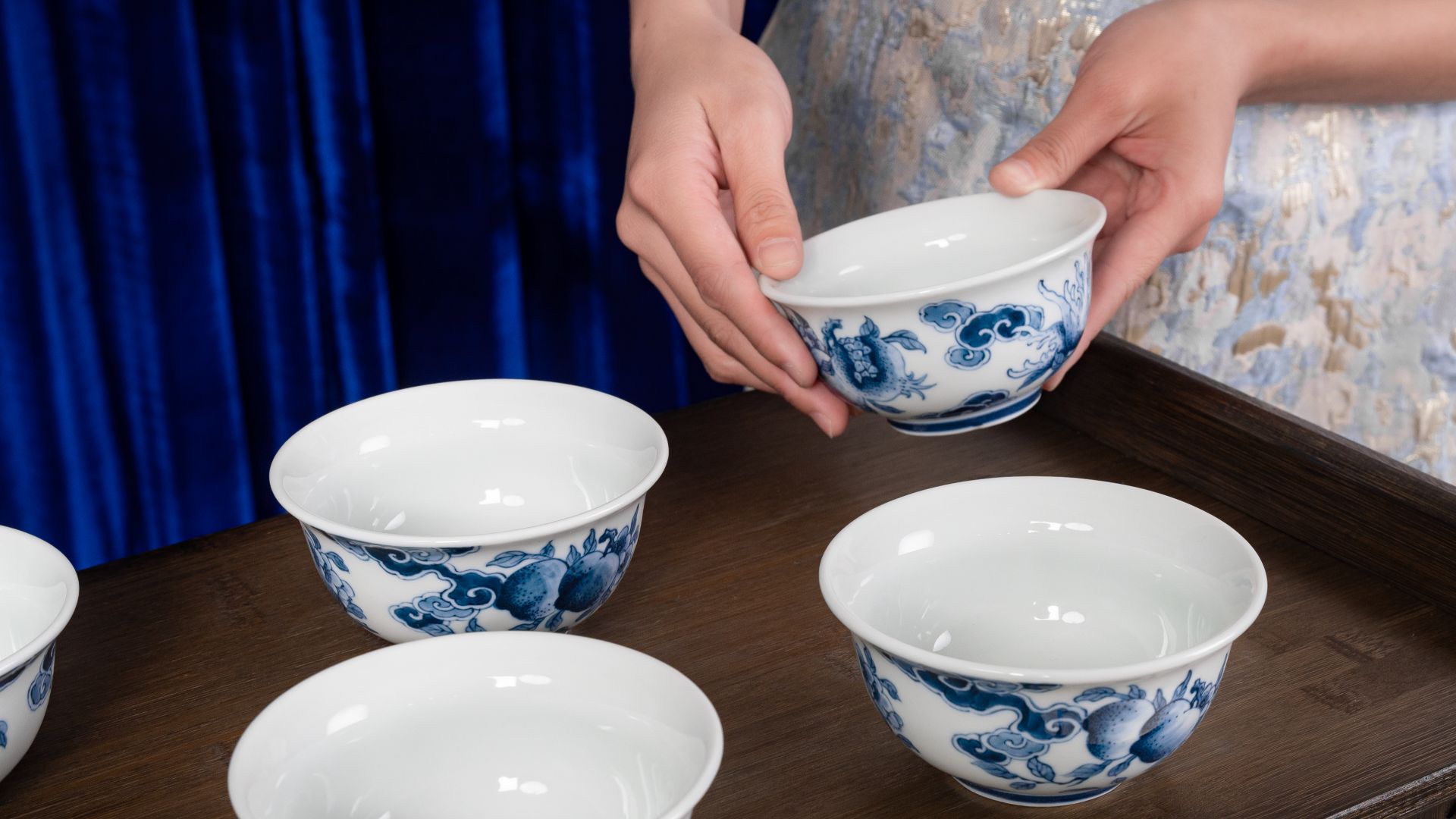 Close-up of elegant blue and white porcelain bowls arranged on a wooden tray with hands carefully placing one.