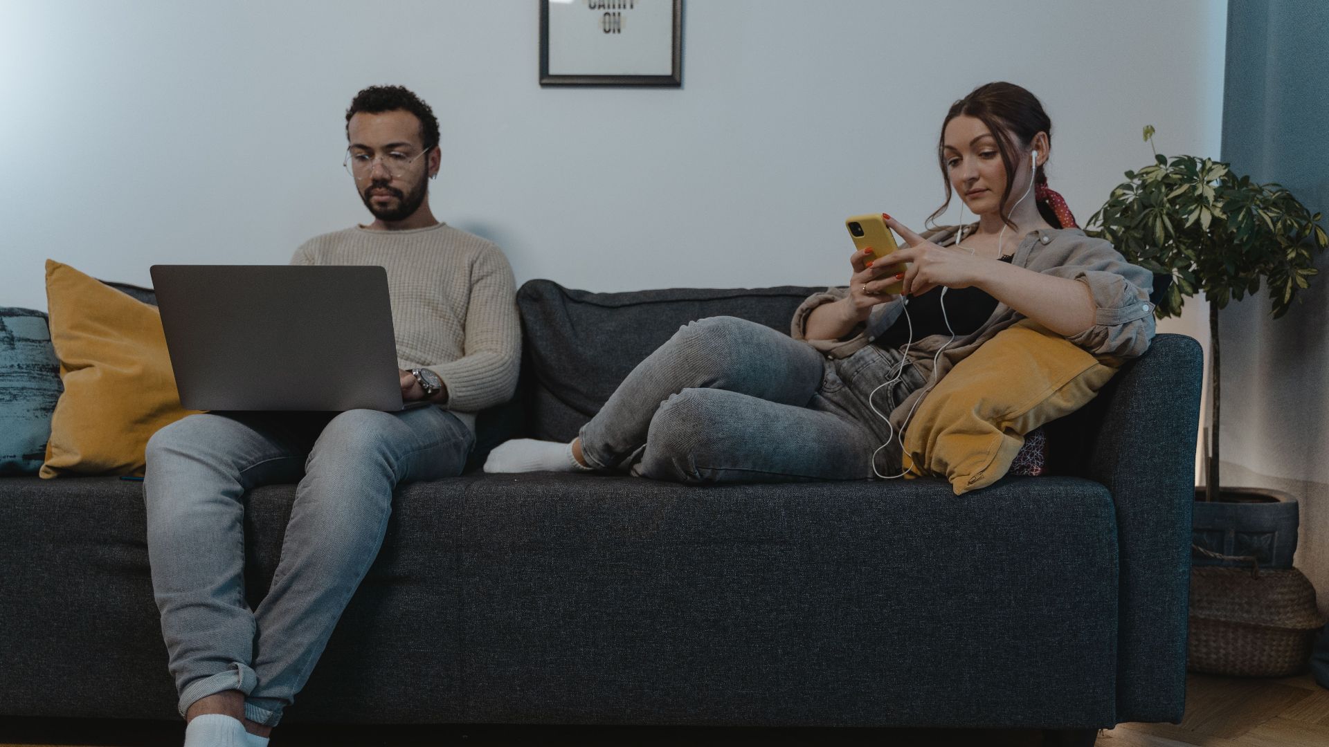 A man and woman relaxing indoors on a sofa, using a laptop and smartphone.