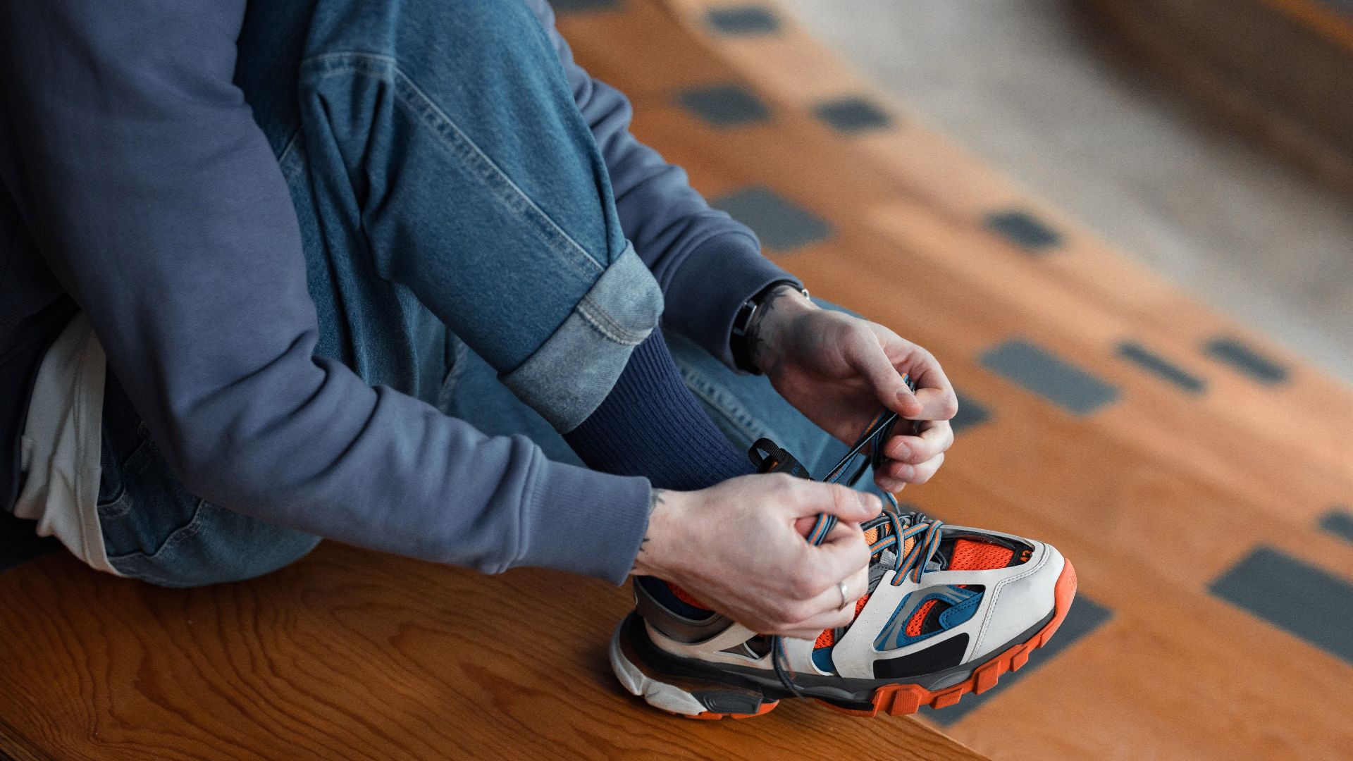 A person tying colorful sneakers on wooden steps, wearing casual denim and sweatshirt.