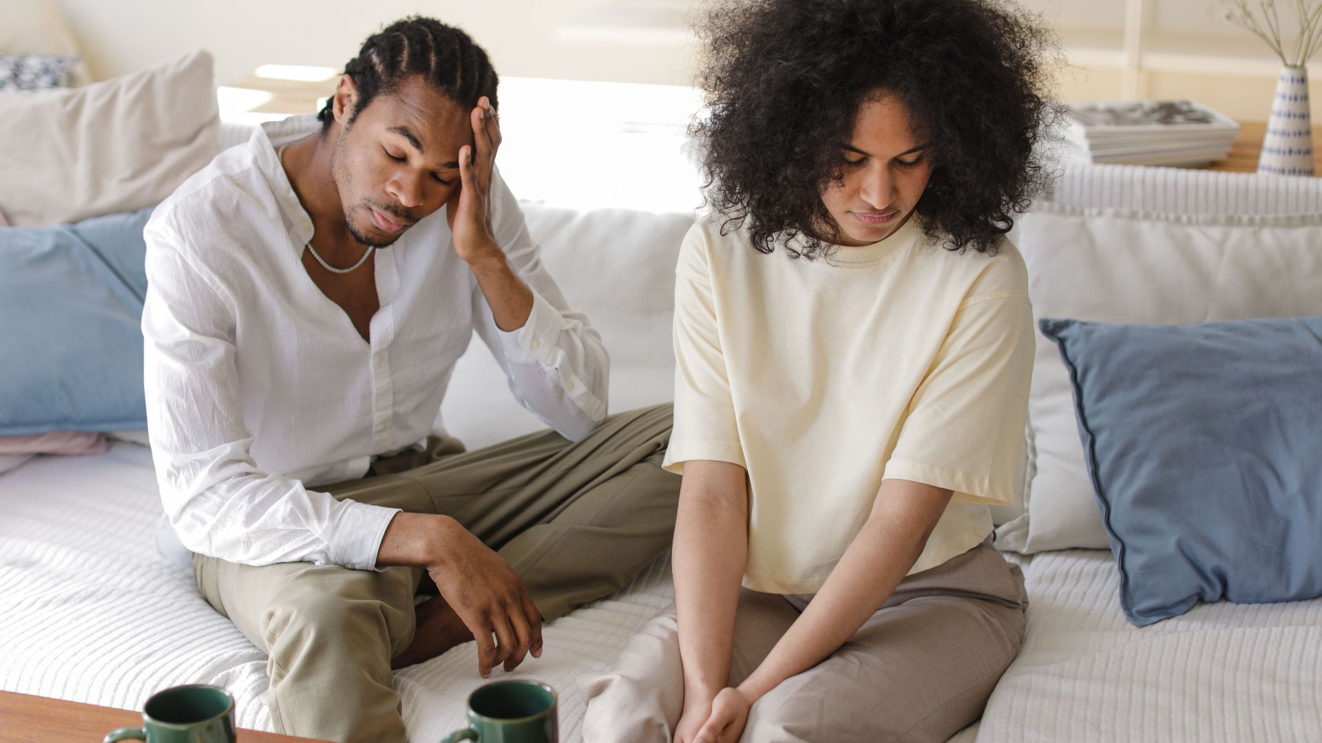 A contemplative black couple sitting on a sofa indoors in a modern setting, reflecting emotions.