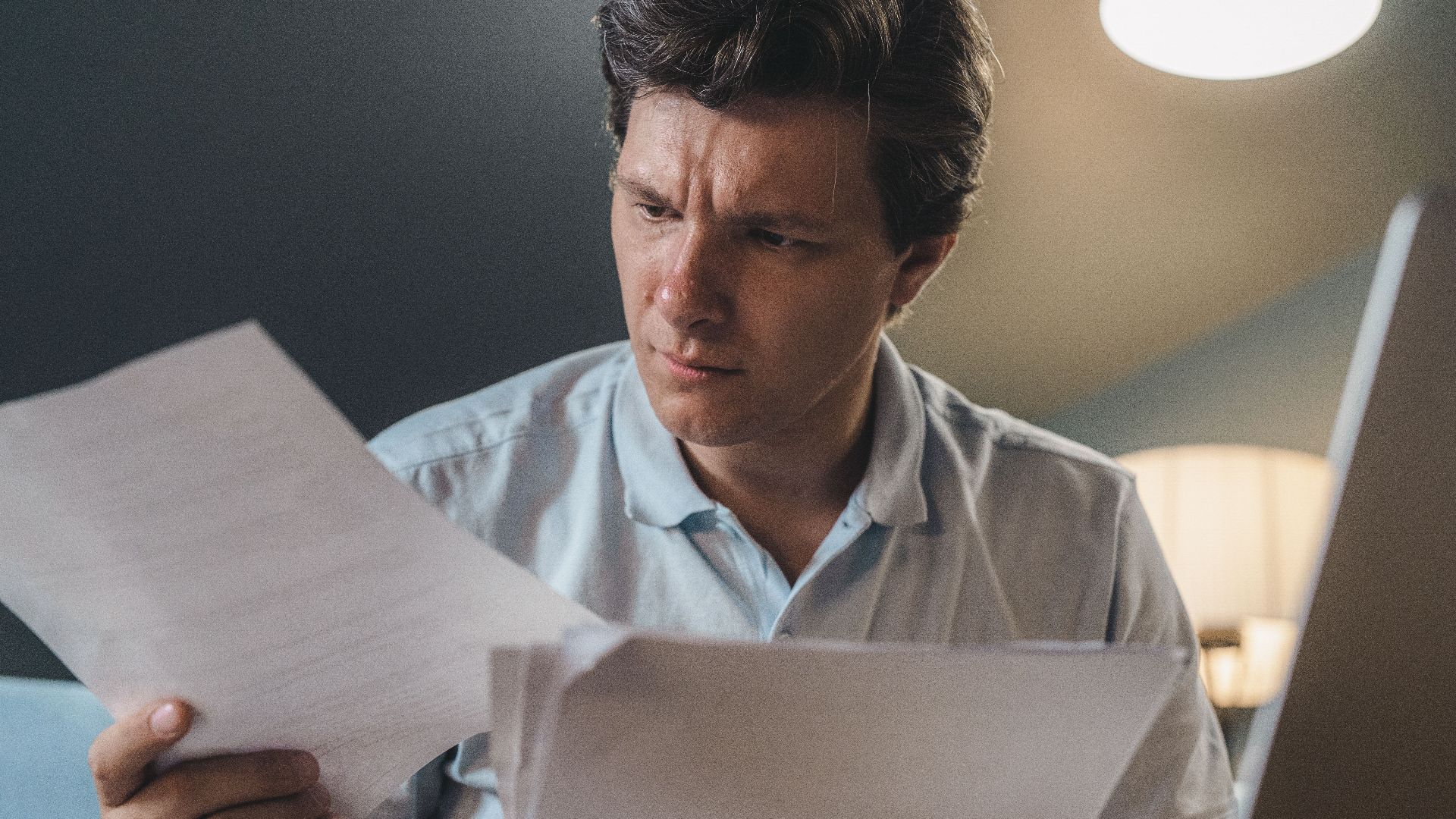 A man intently examines papers, seated indoors under warm lighting, focusing on his work.