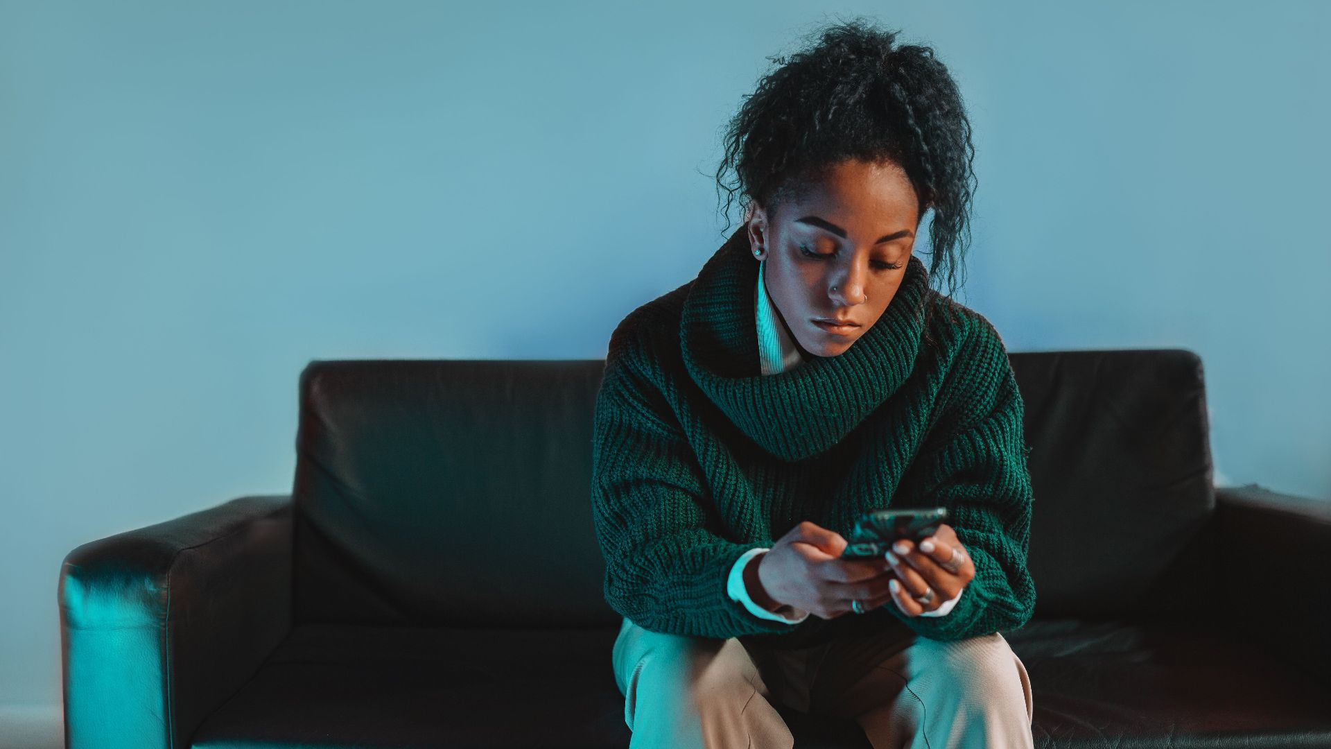 A young woman in a turtleneck sweater focuses intently on her smartphone while sitting indoors.