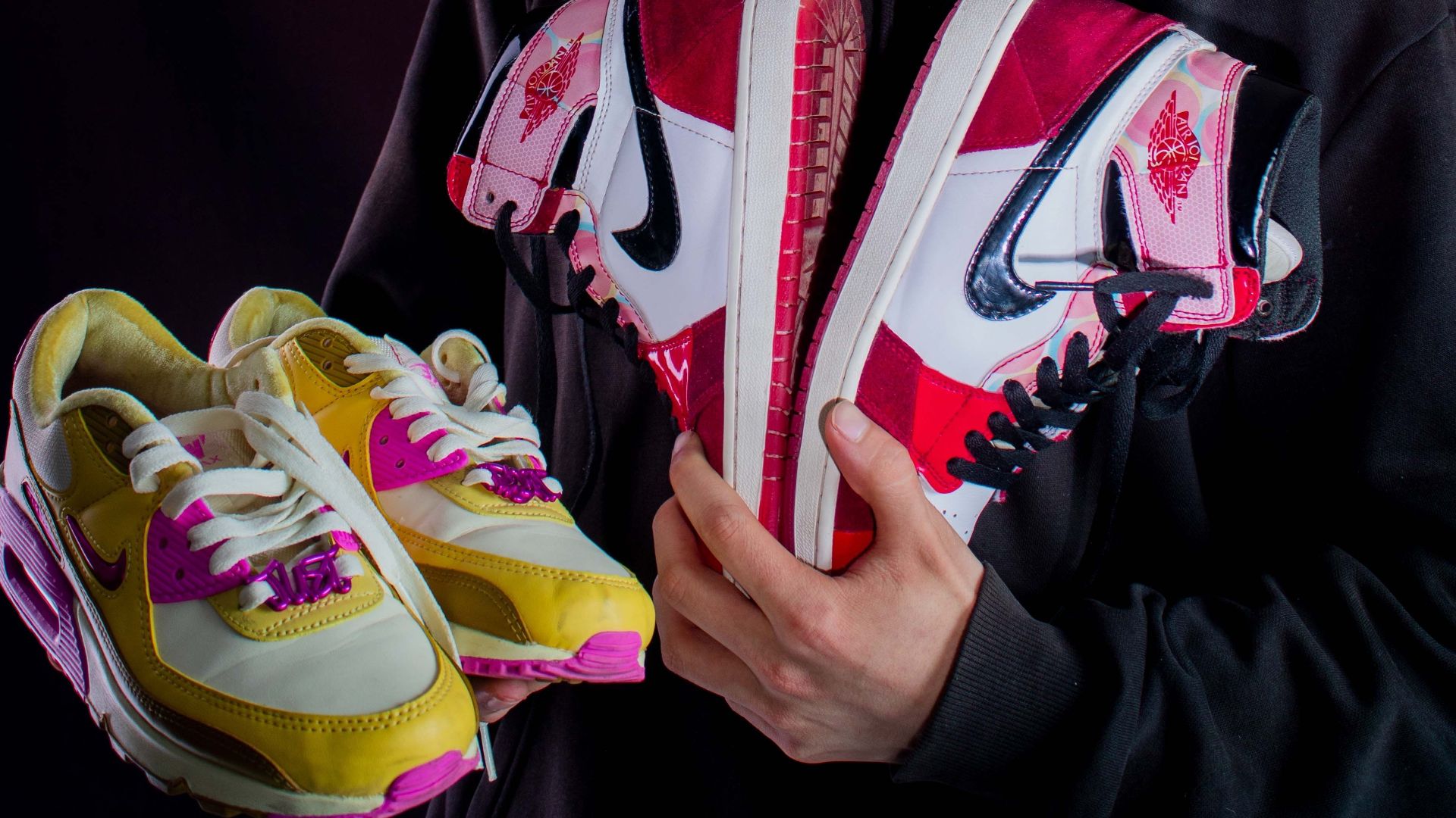 A young man joyfully displays two pairs of vibrant sneakers against a dark backdrop.