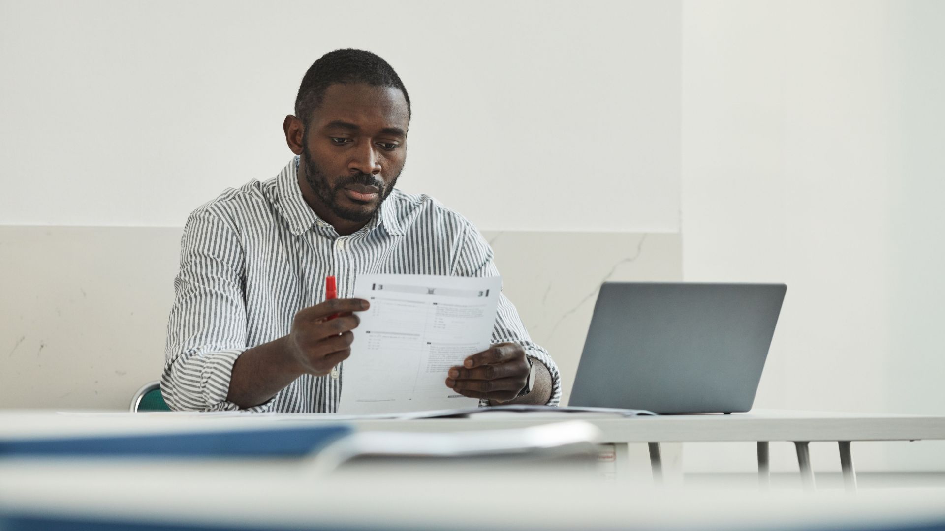 An adult man attentively grading test papers at his workspace with a laptop.
