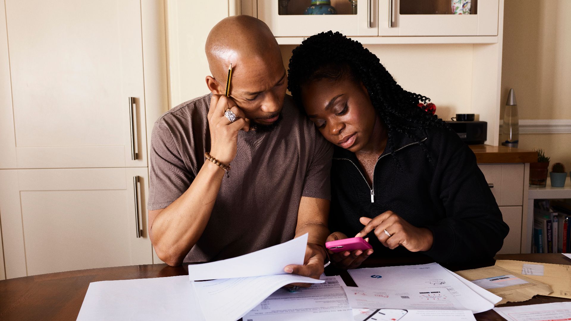 A couple sits at a table managing domestic finances, evaluating documents and using a smartphone.
