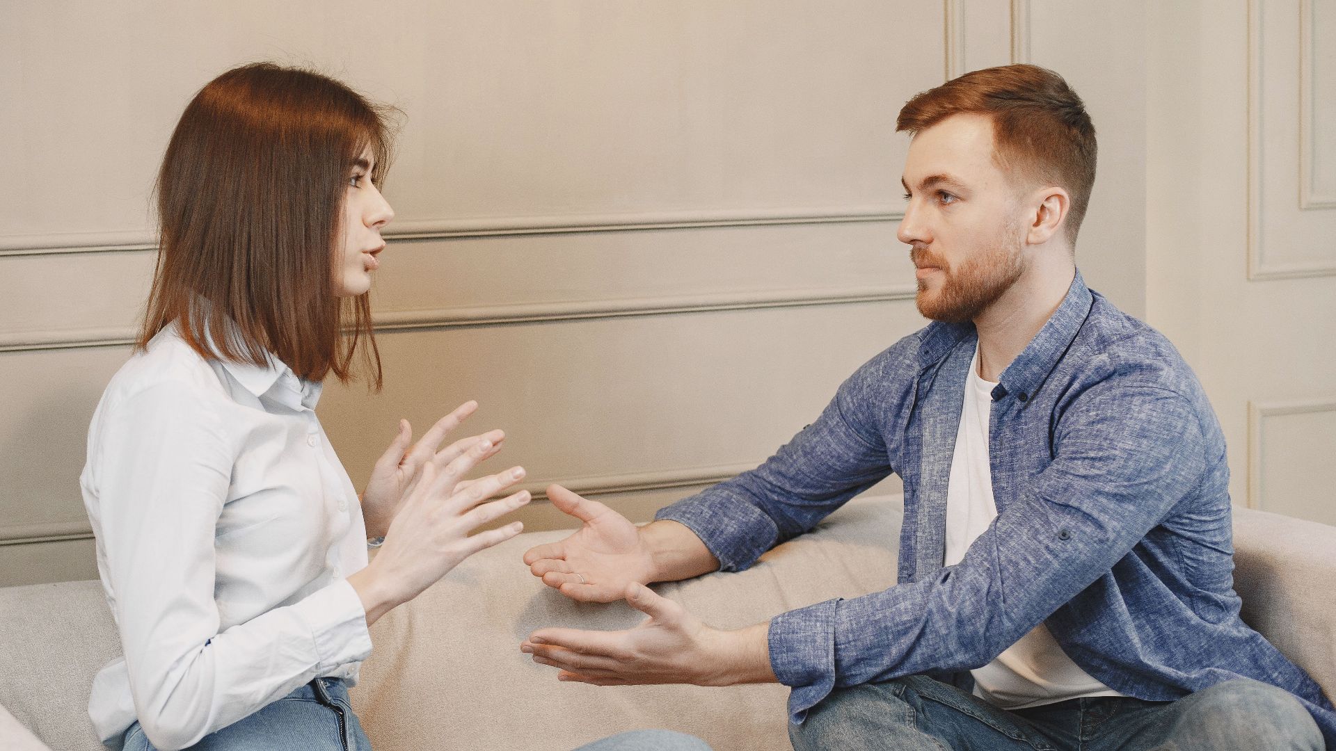 A young couple sitting on a couch having a thoughtful conversation with expressive hand gestures.