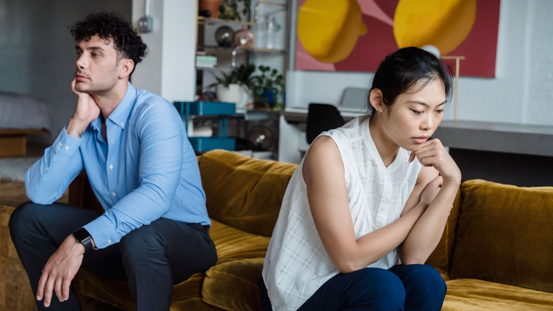 A couple sits indoors on a sofa, both appearing thoughtful and contemplative.