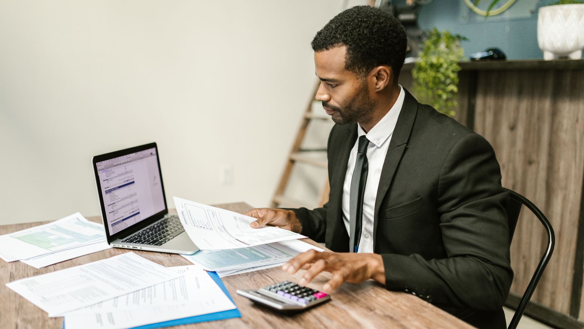 A businessman in a suit working on documents and using a laptop at an office desk.