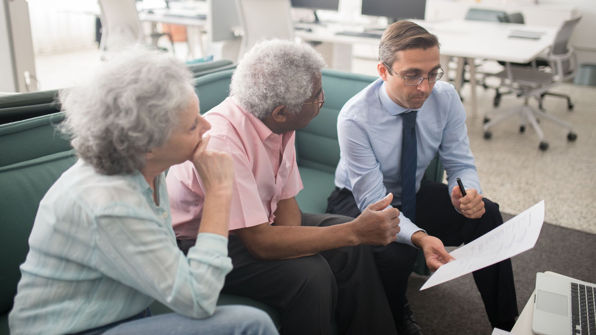 Business professional consults elderly clients in an office setting. Collaborative discussion, paperwork visible.