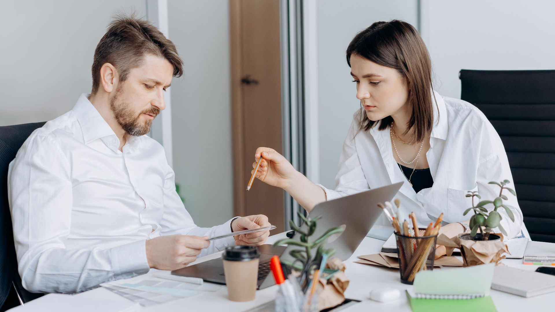 Two colleagues discussing work at a desk with laptops and coffee in a modern office setting.