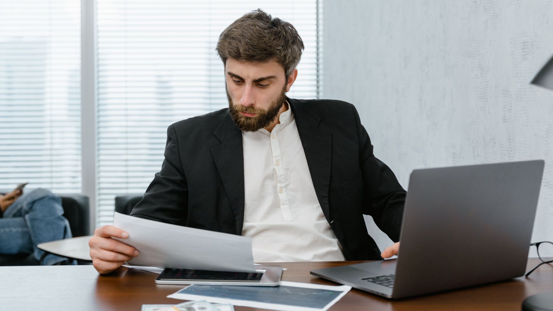 Focused businessman reviews financial reports at office desk with laptop.
