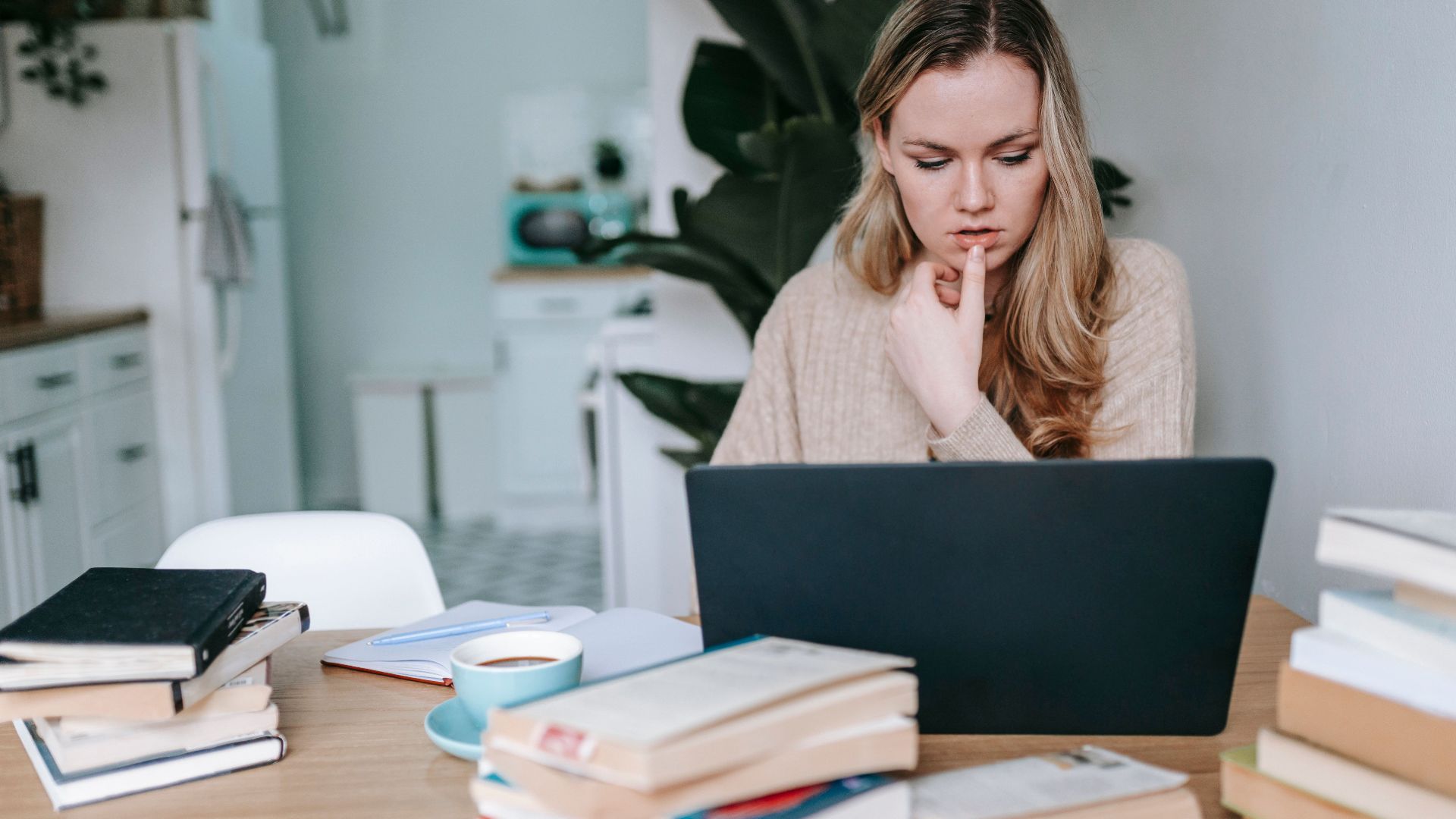 Serious intelligent female studying online on netbook at table with cup of coffee and textbooks on blurred background of home