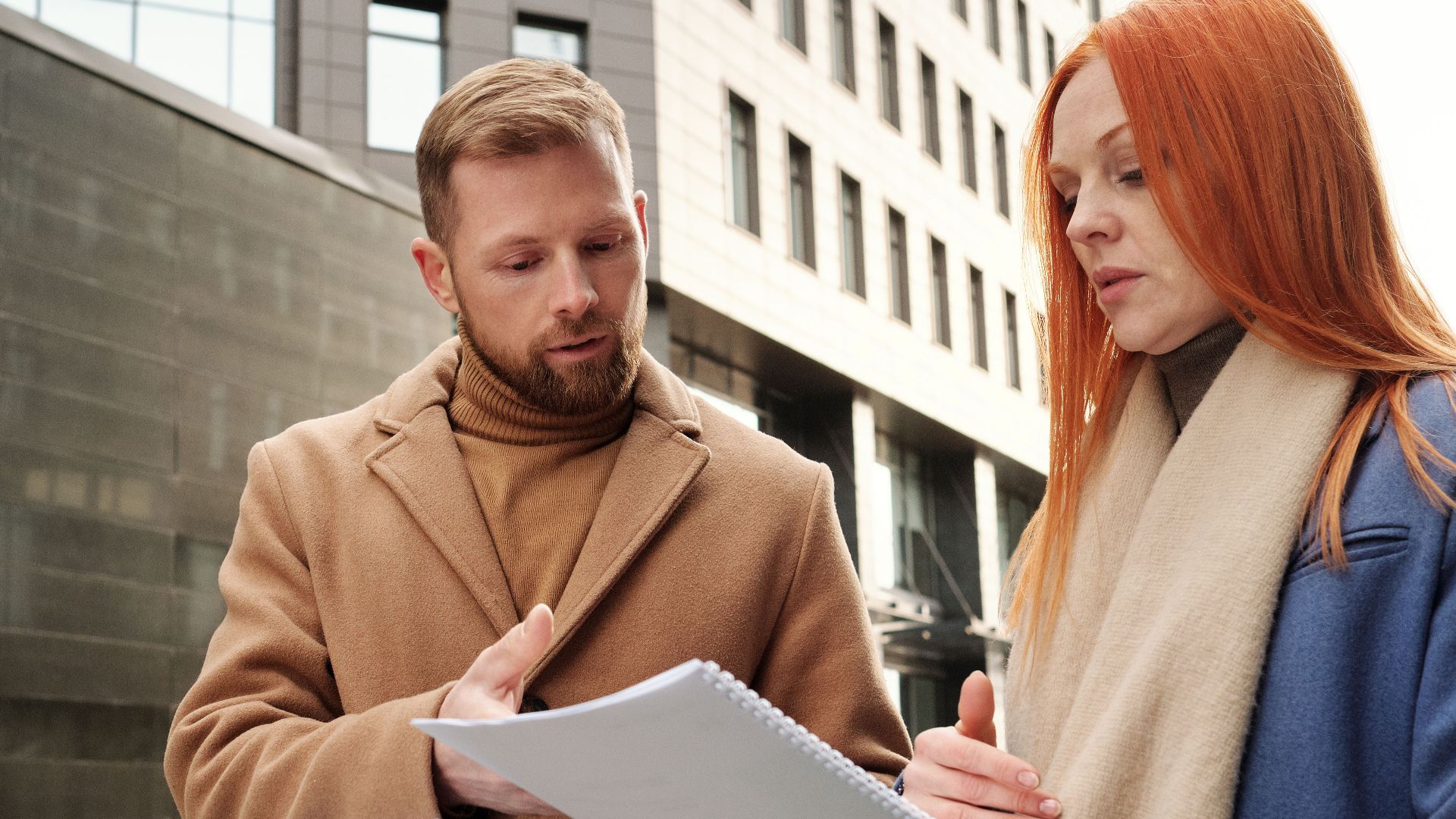 Two business professionals in coats discuss documents in an urban setting.