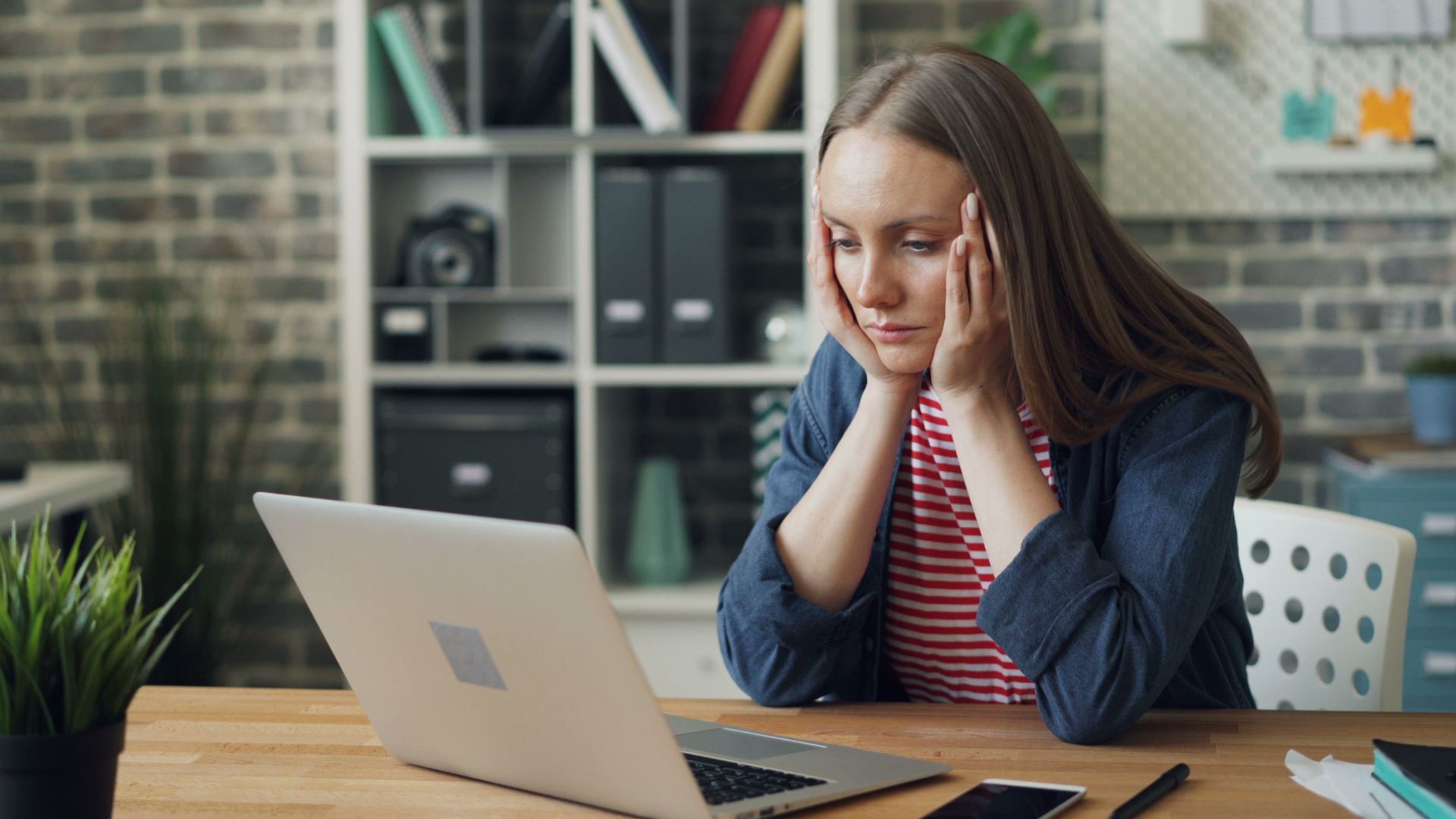 A woman appears stressed while working on a laptop in her modern office.