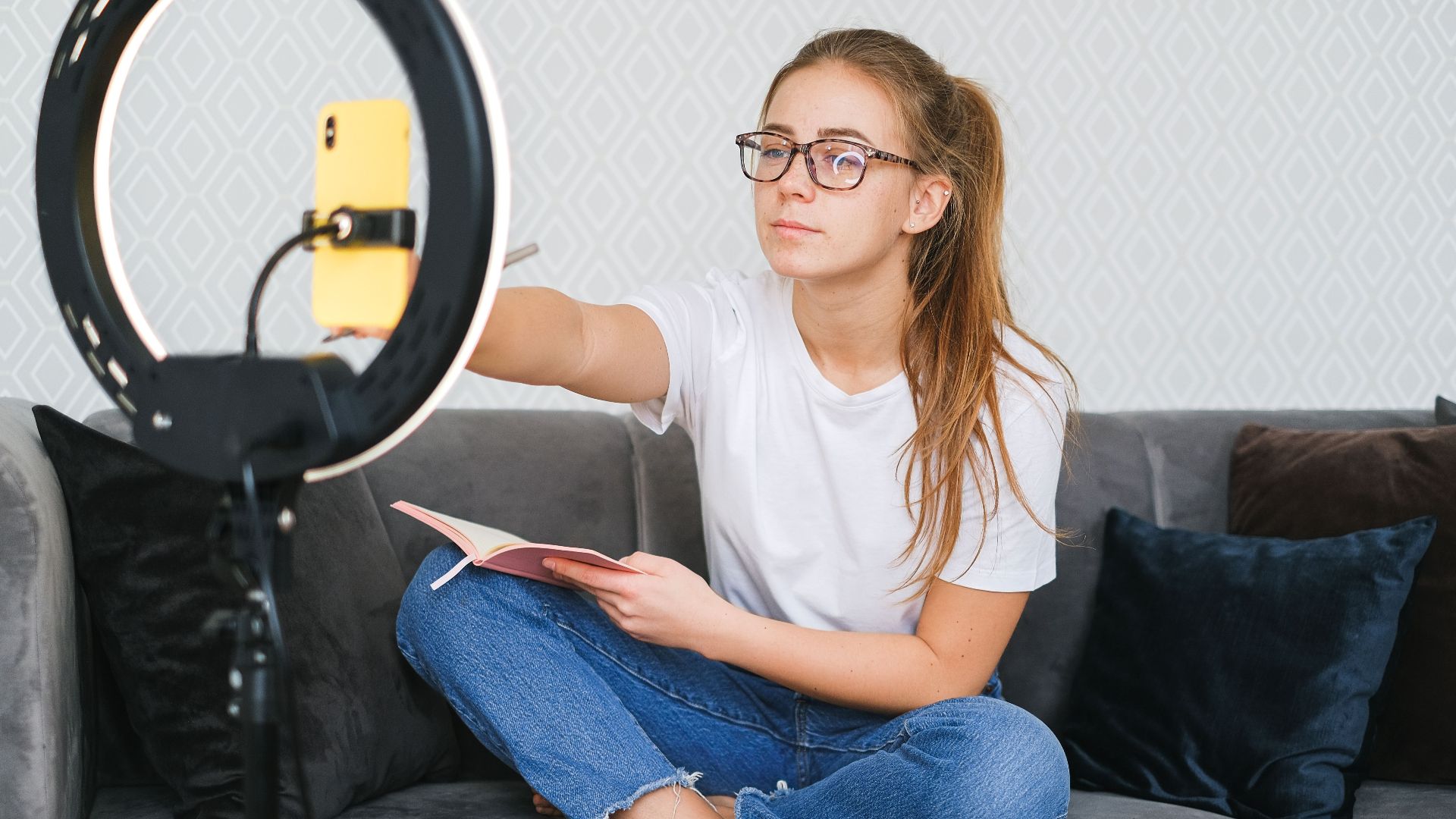 Woman vlogging with smartphone and ring light while holding a book on a sofa.