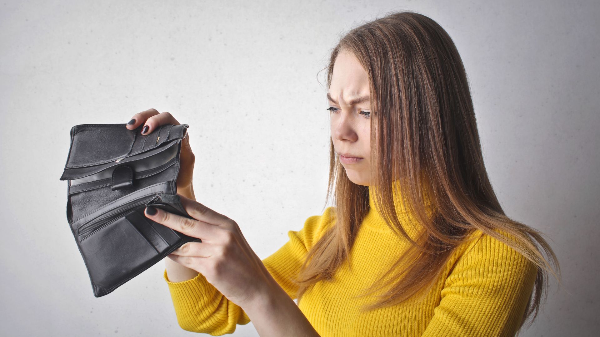 Young woman in a yellow top looking at her empty wallet with a frustrated expression.