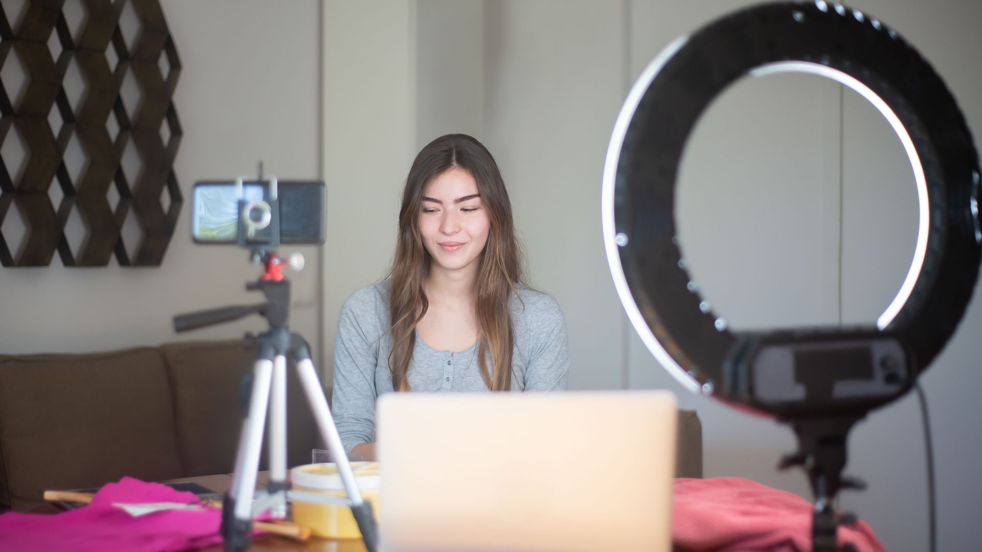 A young woman vlogging indoors using a smartphone, tripod, and ring light setup.
