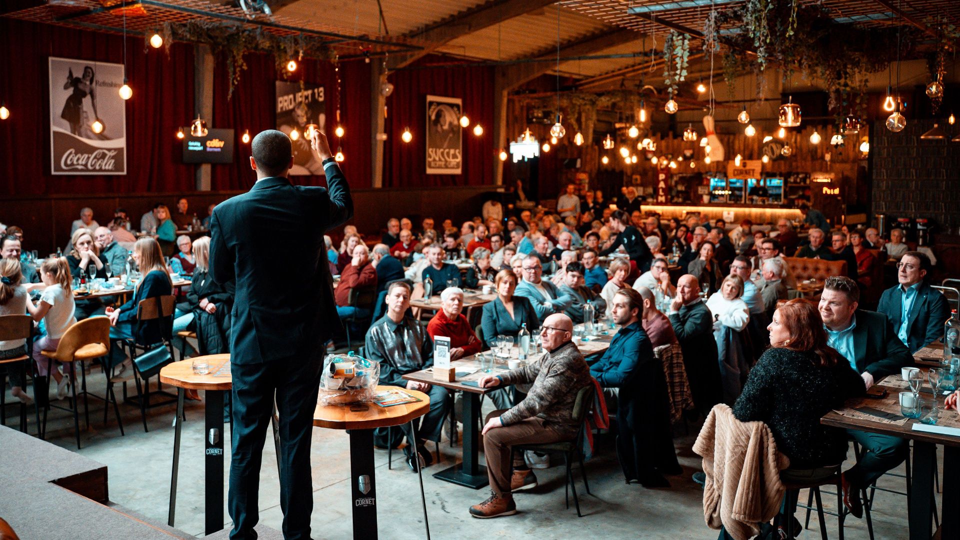 A speaker addressing a large crowd in a vibrant, warmly-lit indoor venue.