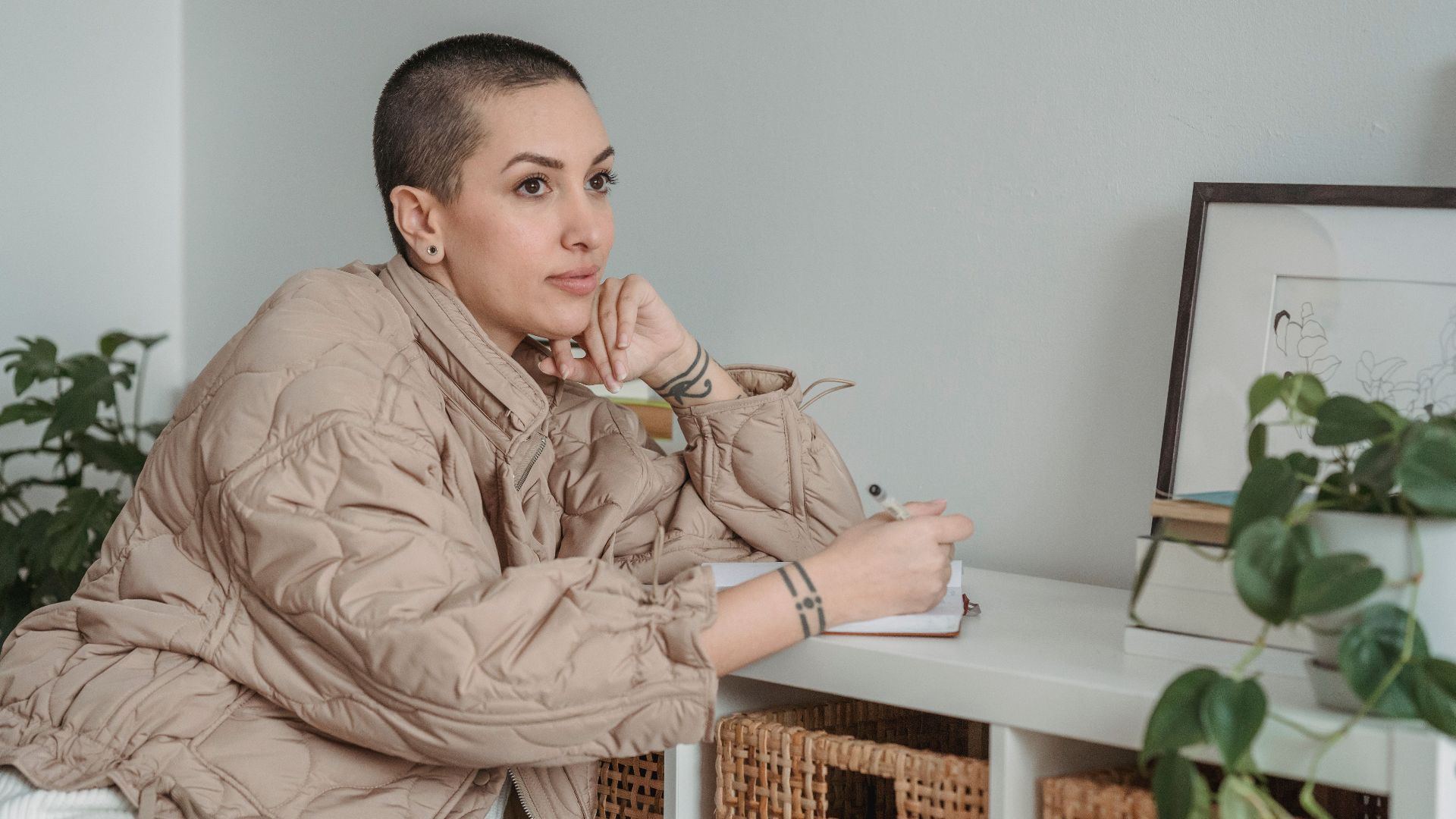 Contemplative female writer with tattoos and short hair leaning on desk with agenda while looking away in house