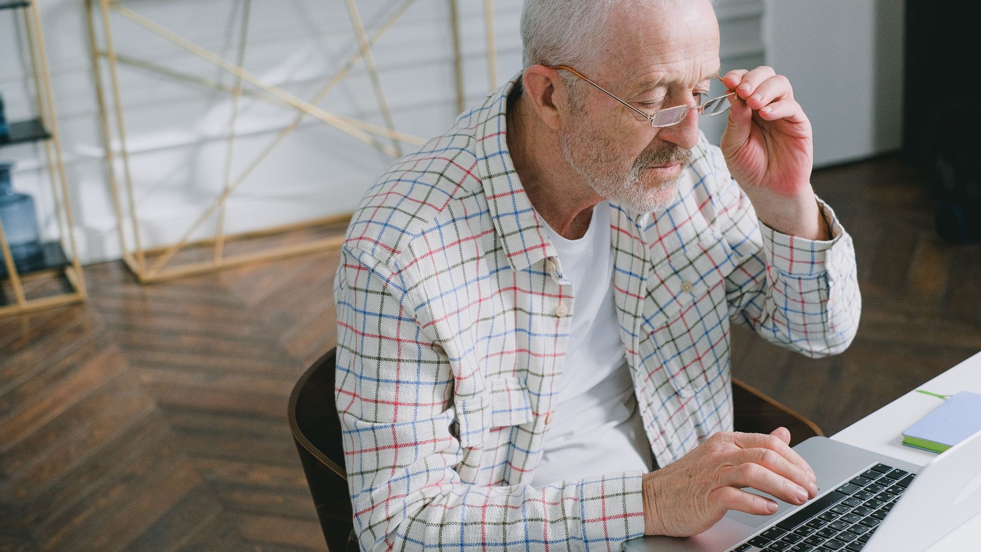 Elderly man wearing glasses focused on working at a laptop indoors.