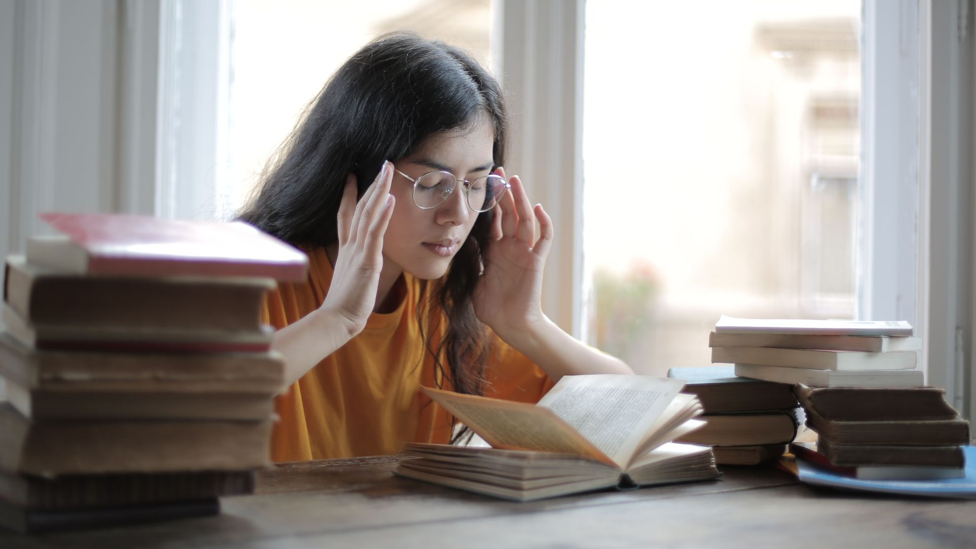 Exhausted ethnic female student in casual clothing sitting at table with stacks of books and suffering from migraine with closed eyes while preparing for exam
