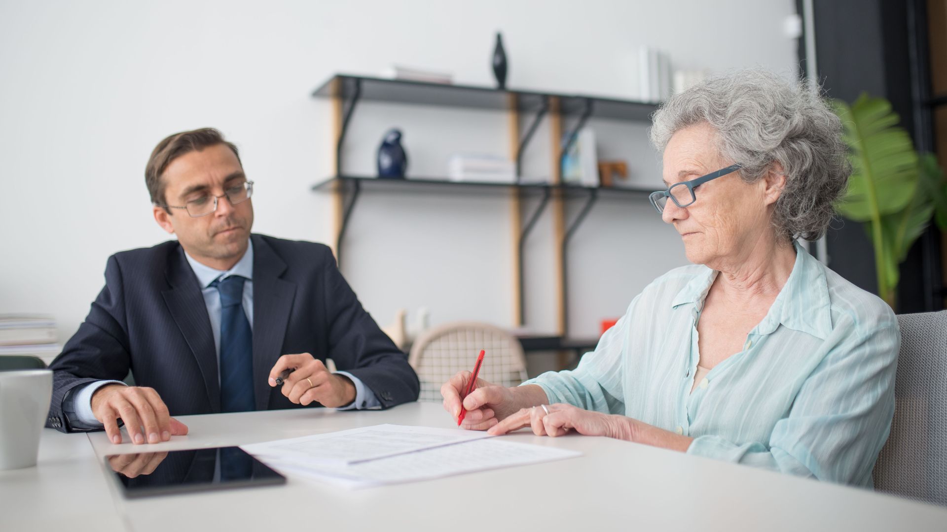 Elderly woman signing paperwork in modern office with consultant.